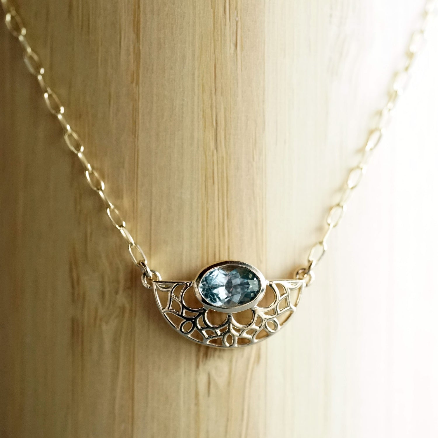 A yellow gold and bezel seat seafoam color sapphire on a light open link chain hanging in front of a bamboo background