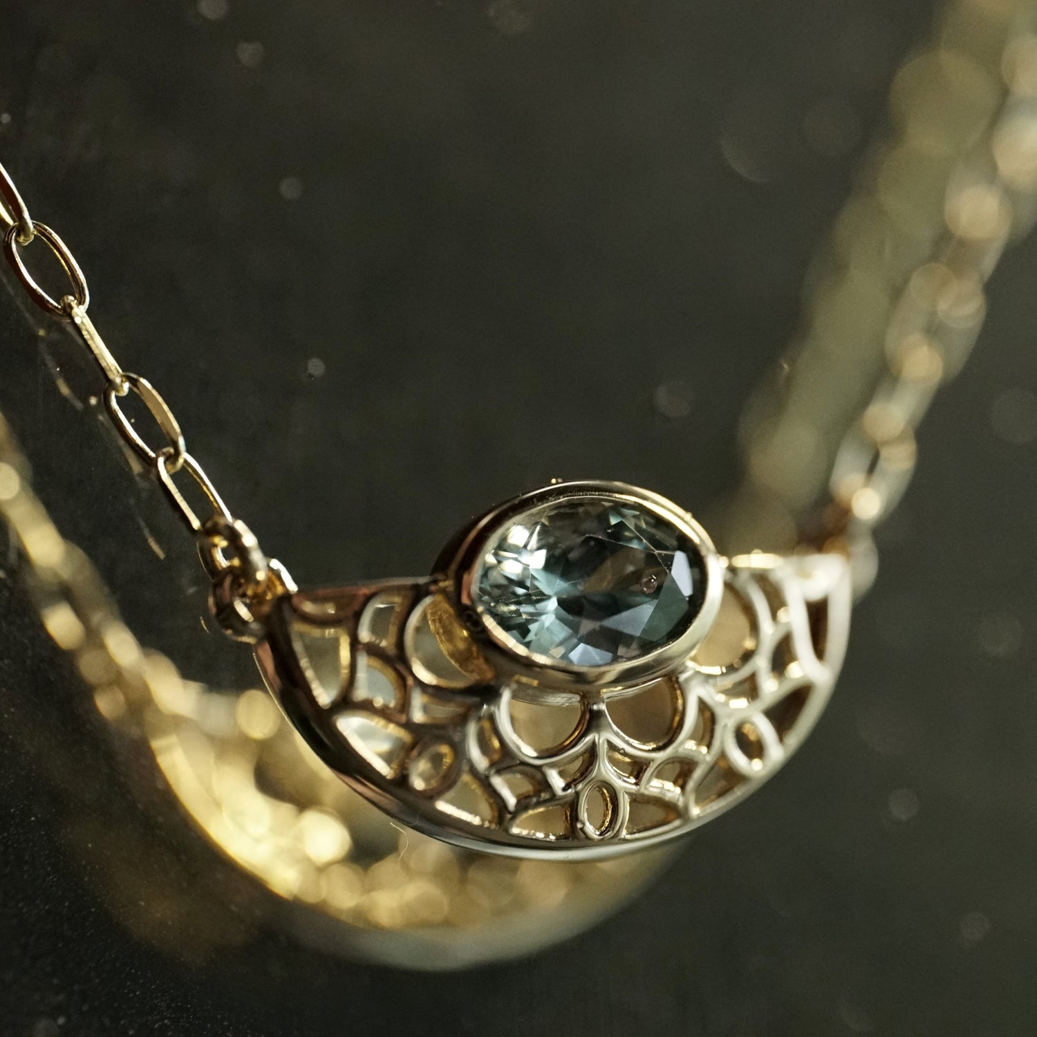 A macro photo of a yellow gold and bezel seat seafoam color sapphire on a light open link chain hanging in front of a black mirrored background