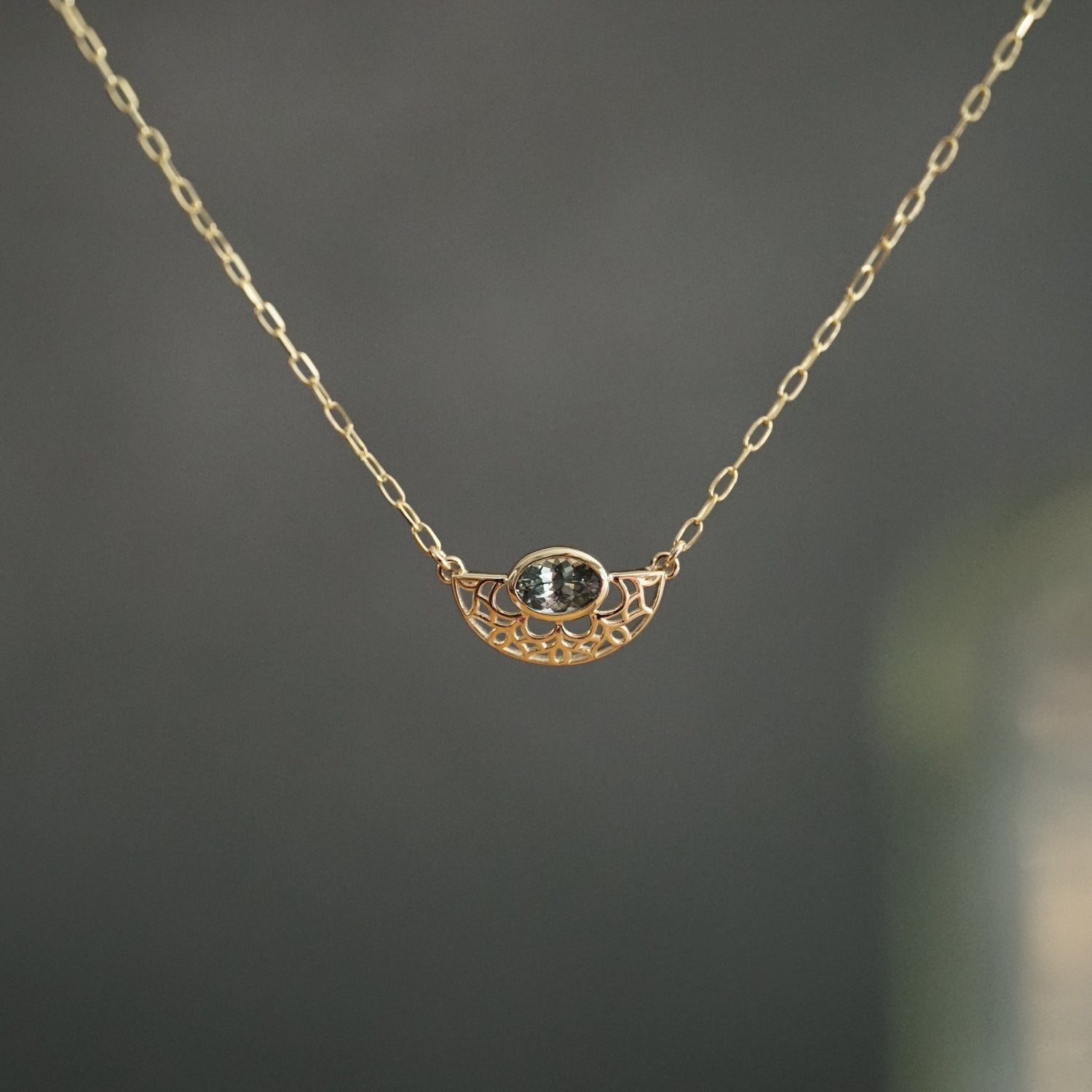 A yellow gold and bezel seat seafoam color sapphire on a light open link chain suspended in front of a grey background