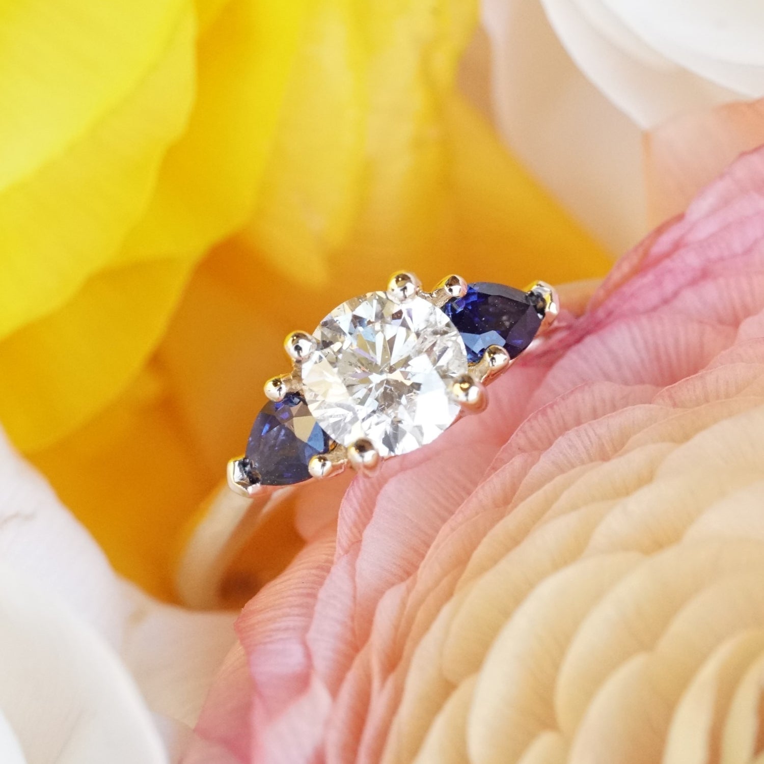 a yellow gold, diamond, and blue sapphire engagement ring rests in yellow white and pink flowers