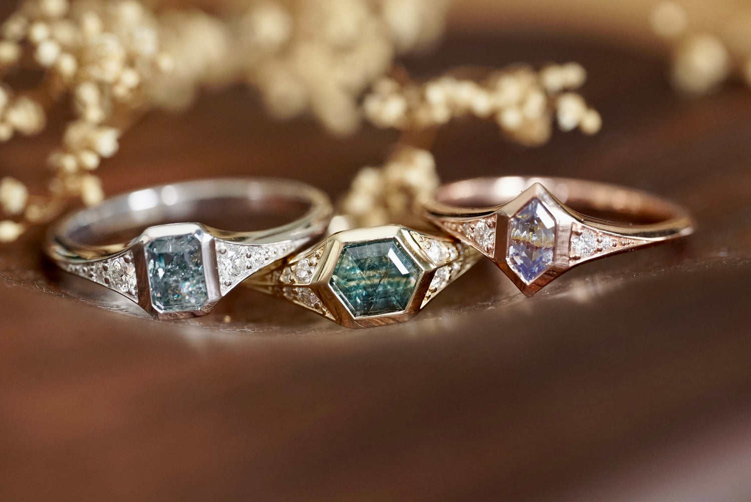 Three sapphire and diamond rings in 14k gold on a wooden surface with flowers in the background