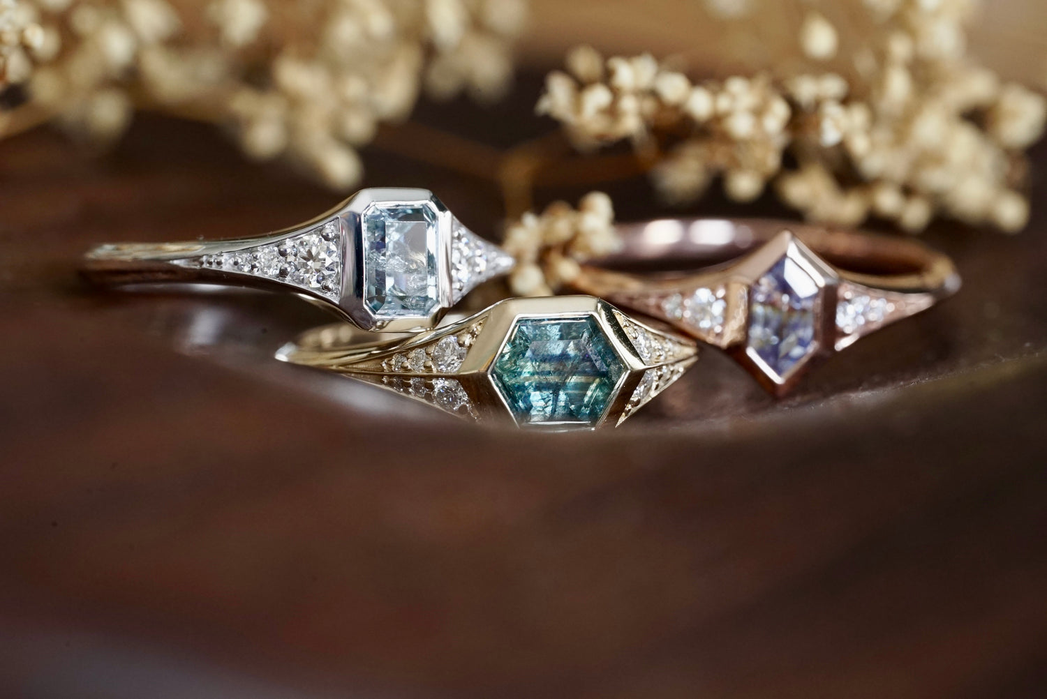 Three sapphire and diamond rings in 14k gold on a wooden surface with flowers in the background