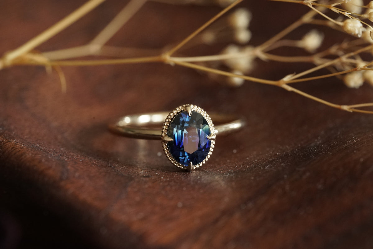A gold ring with an oval blue gemstone resting on a dark wooden surface with dried flowers in the background.