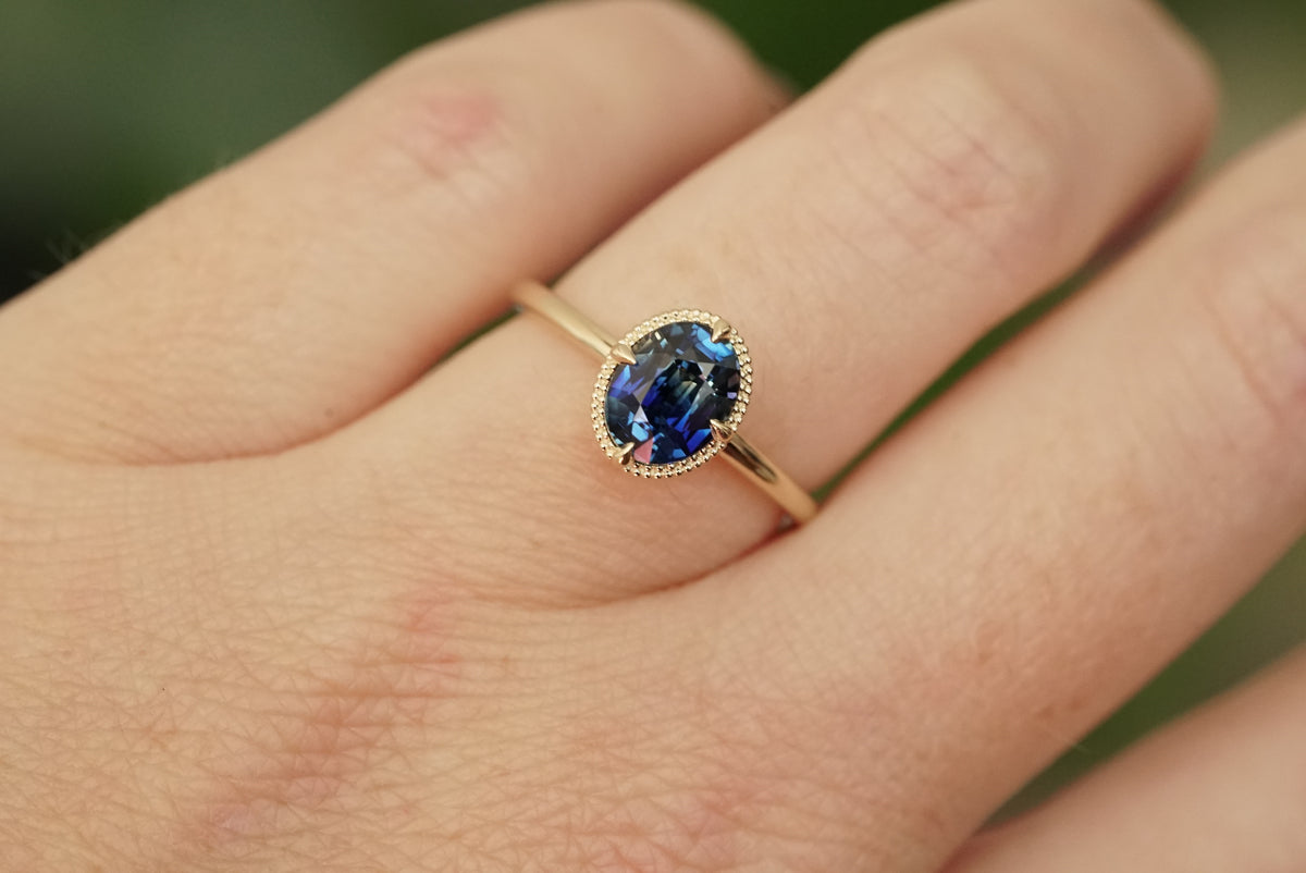 A close-up of a gold ring with a deep blue oval gemstone, worn on a person's finger against a soft, blurred green background.