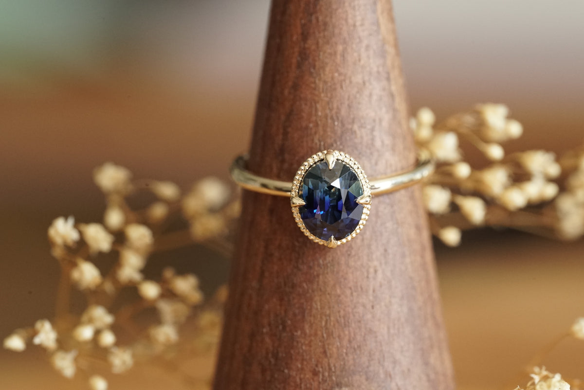 A gold ring with an oval-cut dark blue gemstone, displayed on a wooden cone stand with dried flowers in the background.