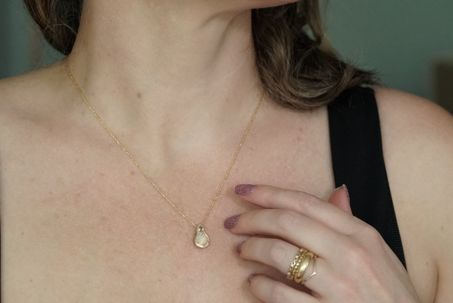 Close-up of a person wearing a gold necklace with a teardrop pendant and multiple gold rings on a neutral background.