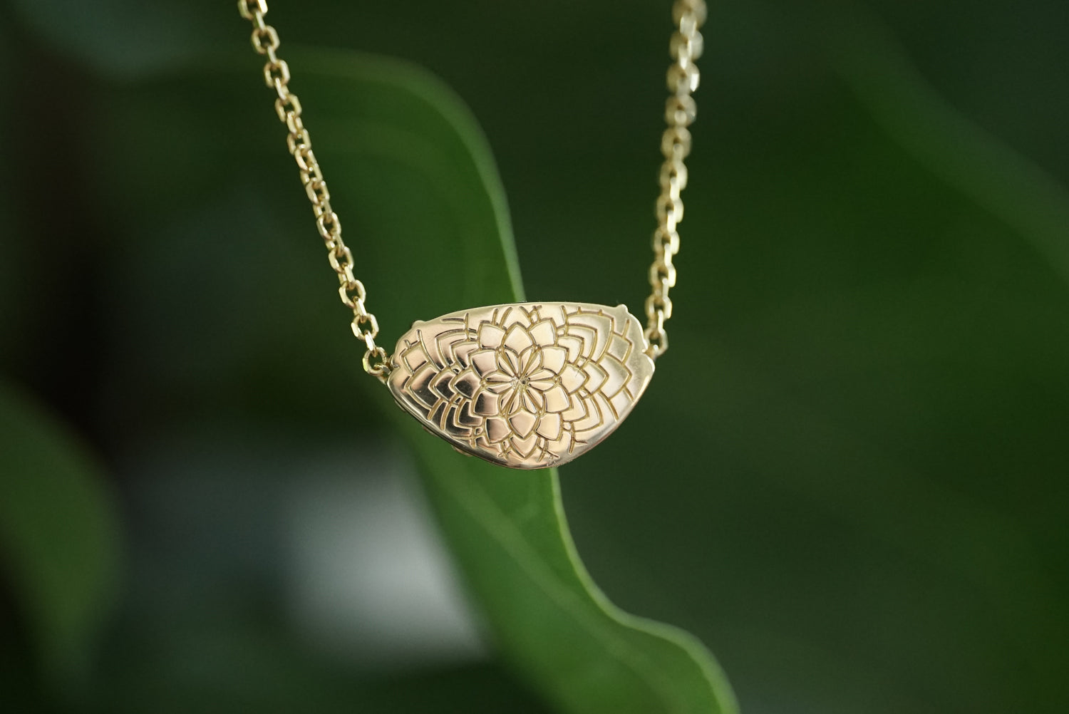 A gold pendant necklace with an intricate floral mandala pattern, hanging against a blurred green leaf background.