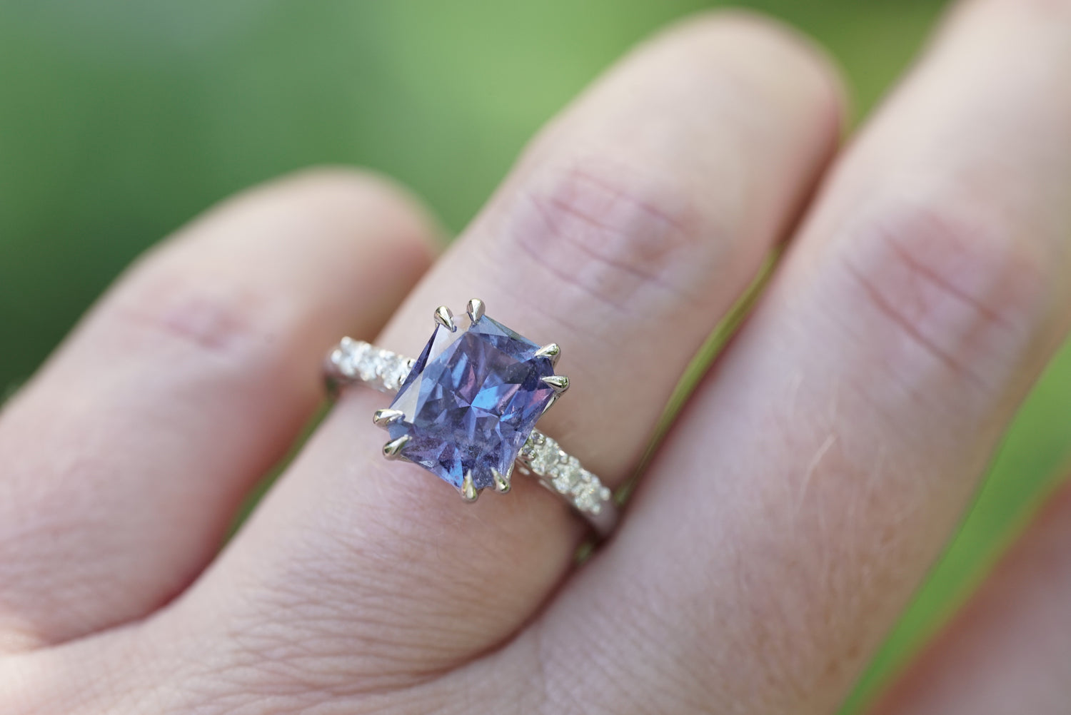 A close-up of a hand wearing a platinum ring with a large rectangular purple sapphire and small diamond accent stones along the band, set against a blurred green background, taken in bright sunlight outside