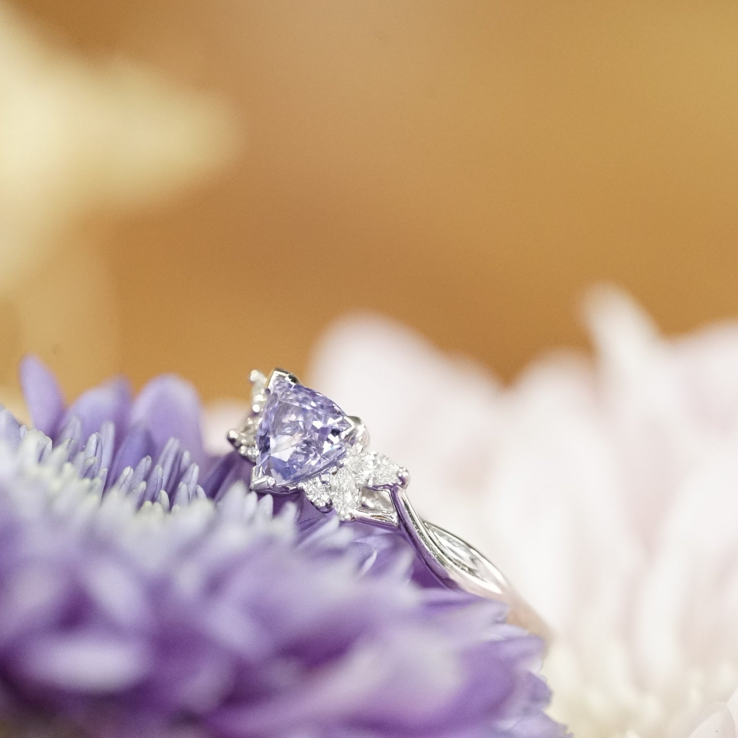 A macro photo of a triangular shaped lavender spinel and diamonds set in a white gold ring laying on a purple flower with a yellow backdrop