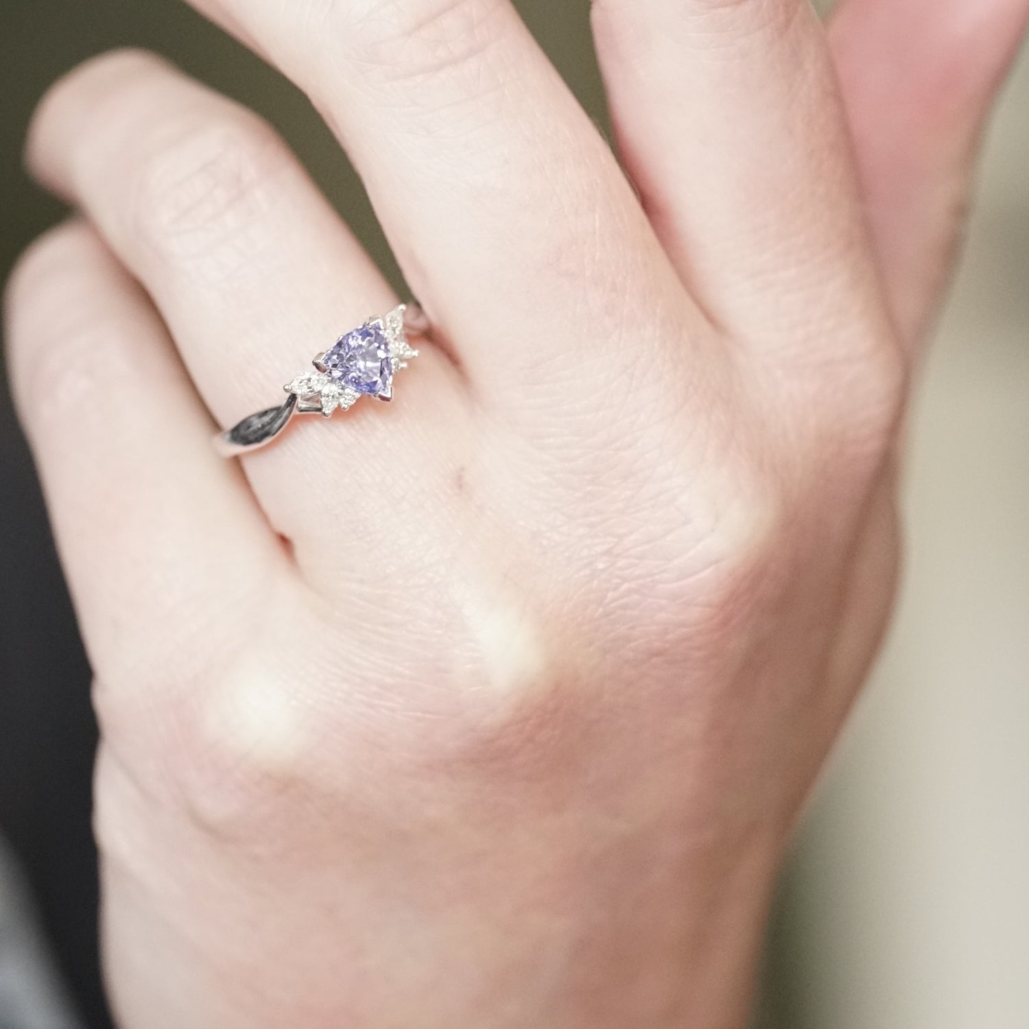 A photo of a triangular shaped lavender spinel and diamonds set in a white gold ring on a light skinned left hand