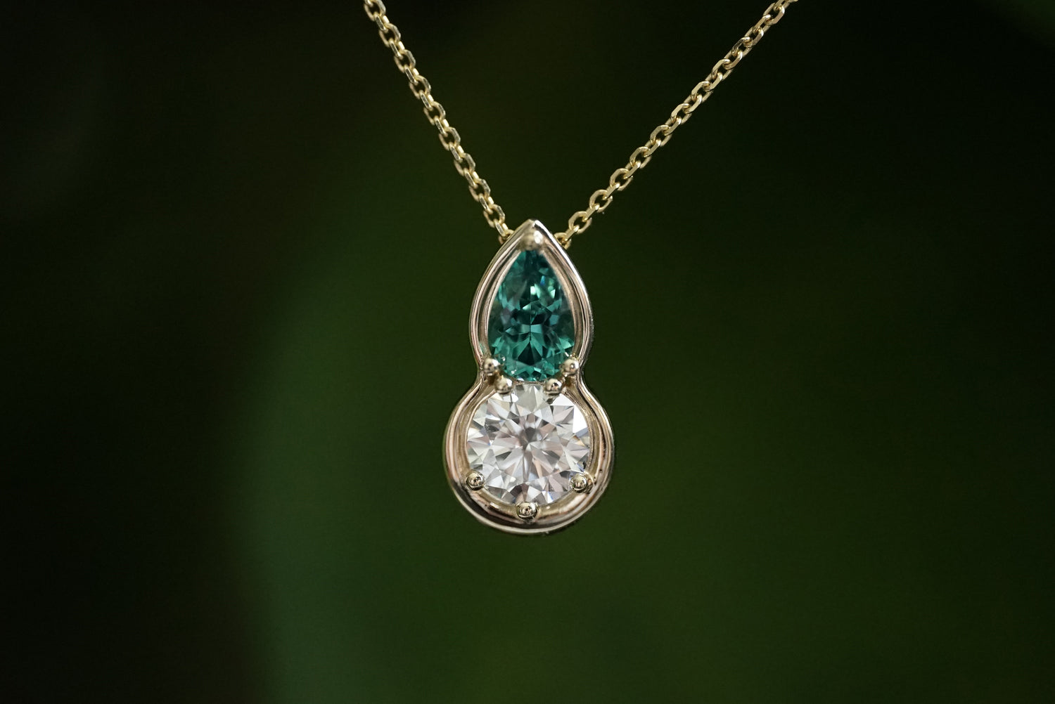 Teardrop-shaped pendant with green and clear gemstones on a gold chain against a dark background