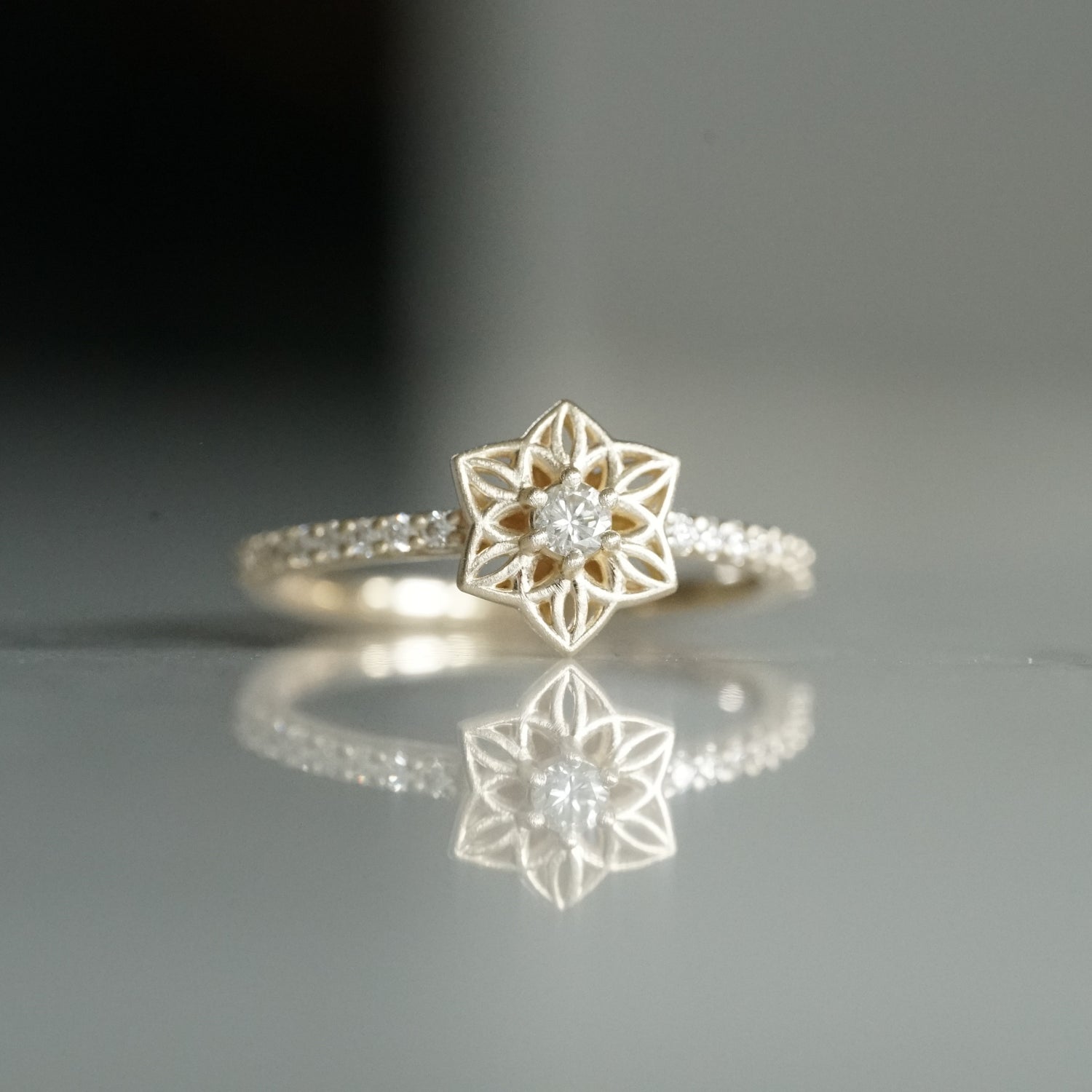 a yellow gold ring with an open wirework flower inspired by daffodils with a single diamond prong set center stone and shared prong diamonds on the shank on a reflective background