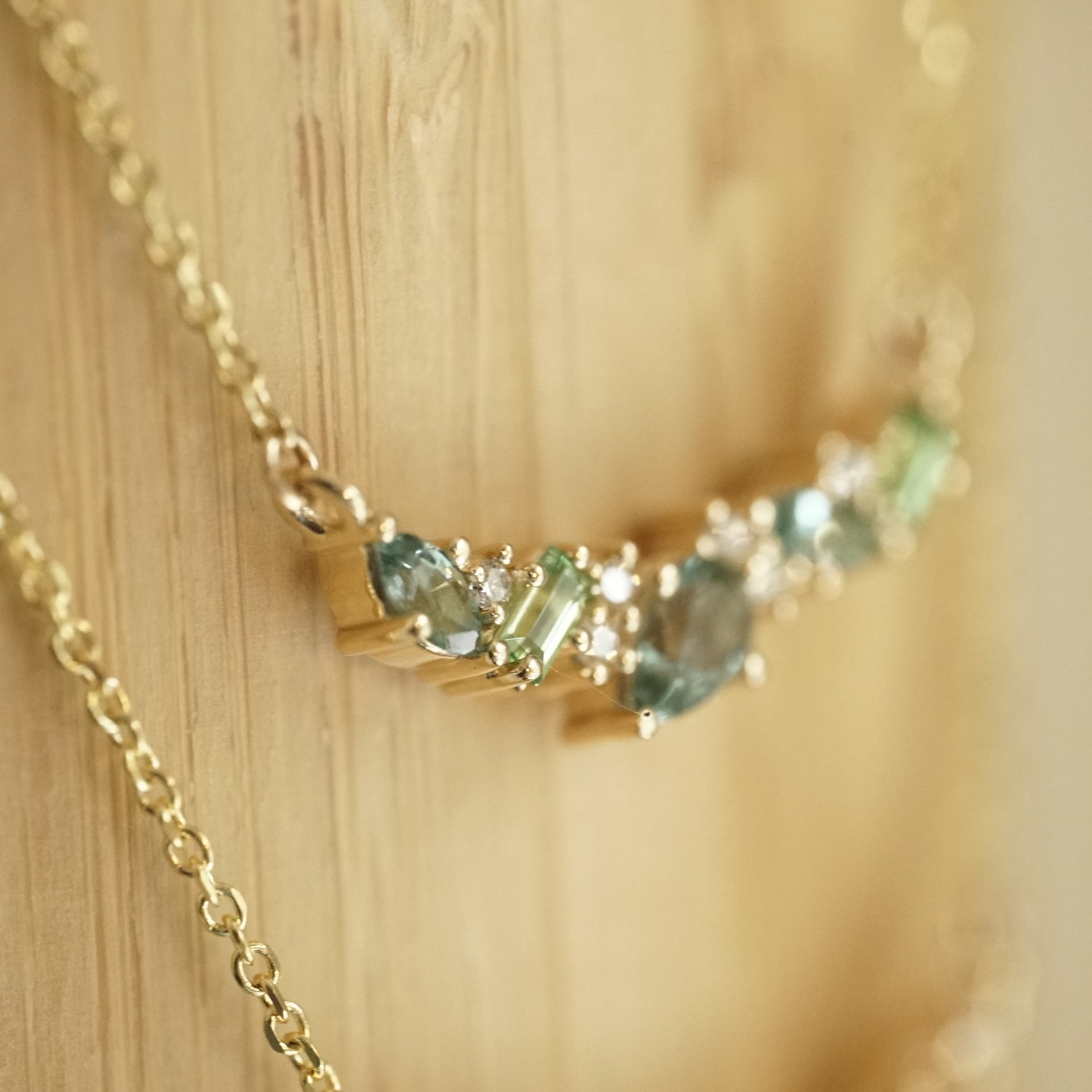 a macro photo of a yellow gold pendant features green and teal gemstones paired with round white diamonds in a playful design hangs on an open link chain in front of a bamboo backdrop