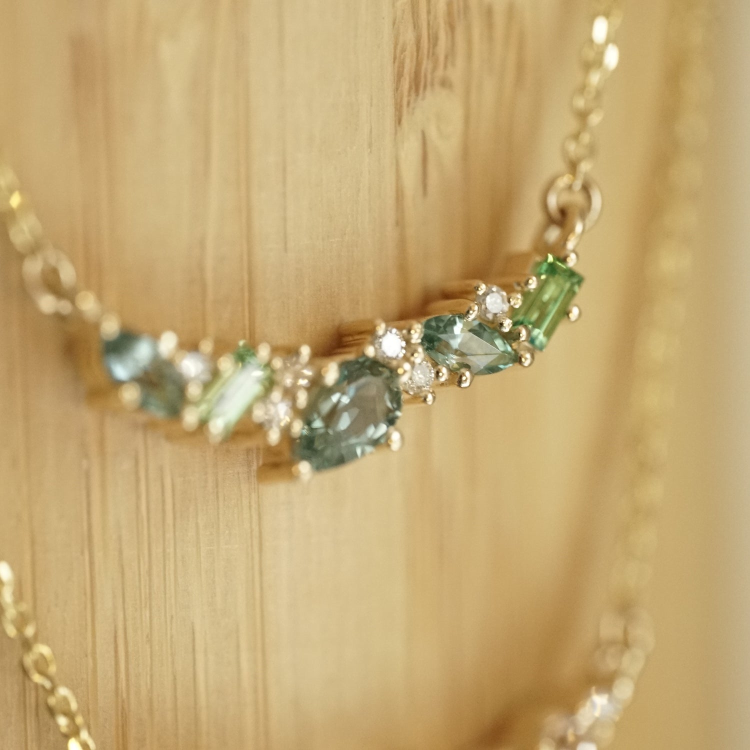 a macro photo of a yellow gold pendant features green and teal gemstones paired with round white diamonds in a playful design hangs on an open link chain in front of a bamboo backdrop