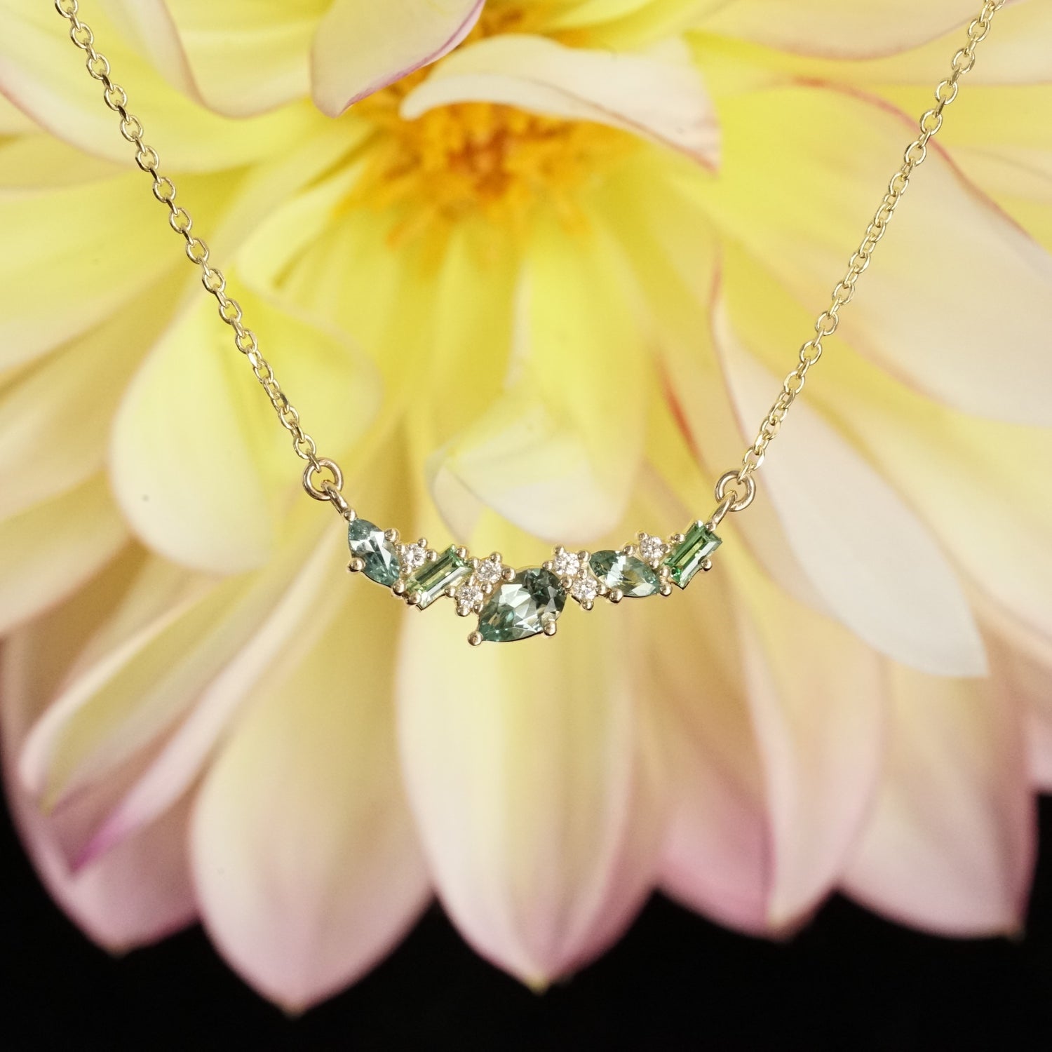 a yellow gold pendant features green and teal gemstones paired with round white diamonds in a playful design hangs on an open link chain in front of a pink and yellow flower