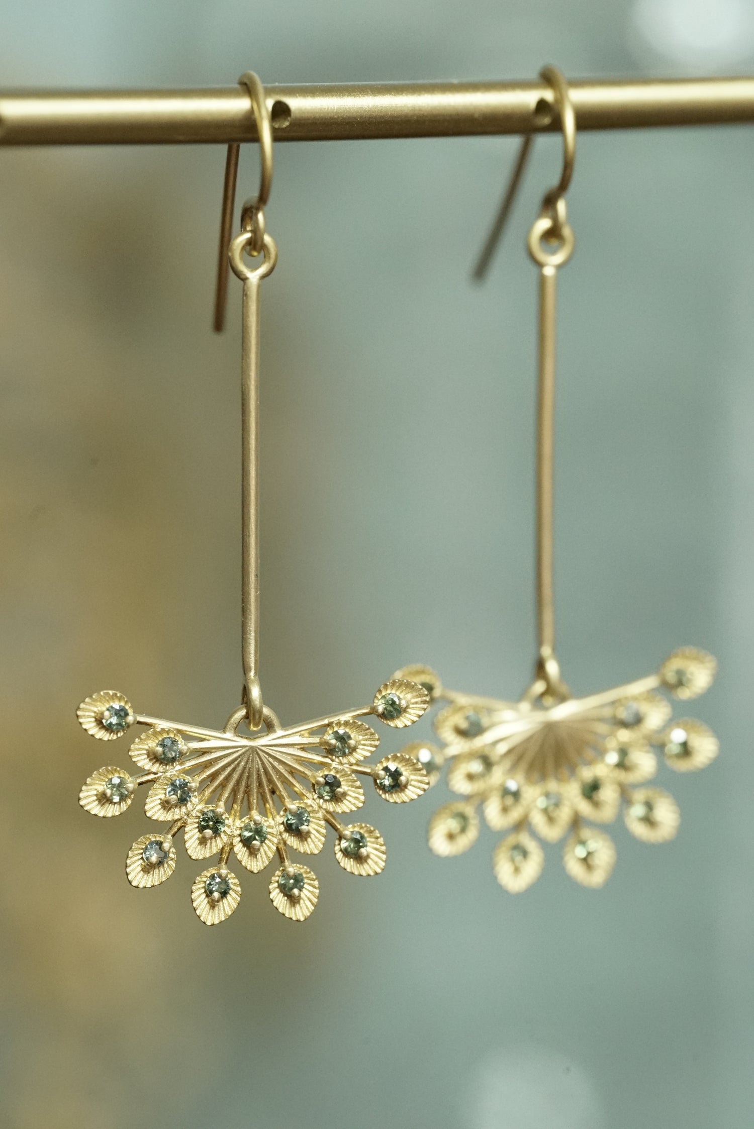a pair of 14k yellow gold and teal sapphire drop earrings hangs on a brass bar