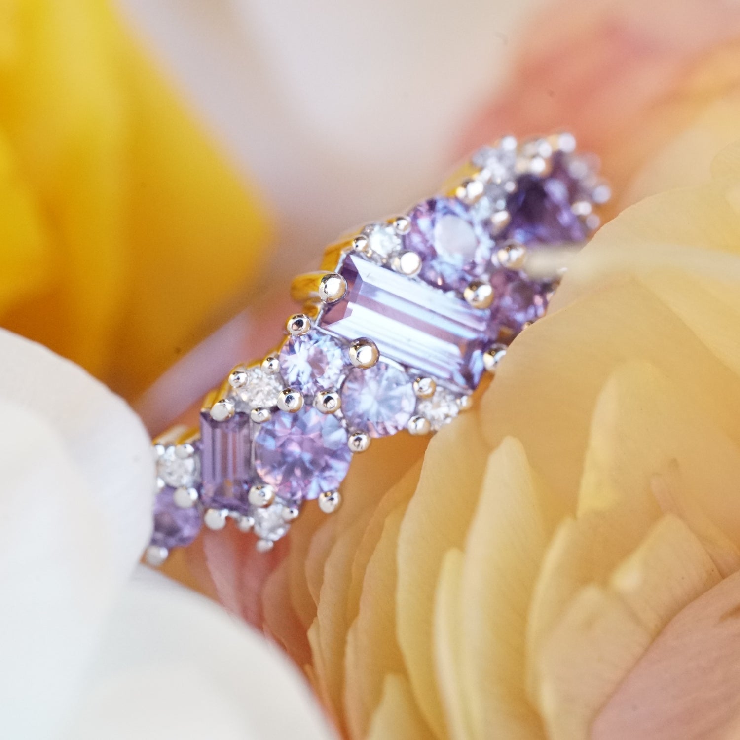 purple sapphires of mixed sizes and shapes are set in a white gold ring and accented with diamonds. the ring is laying on a yellow flower