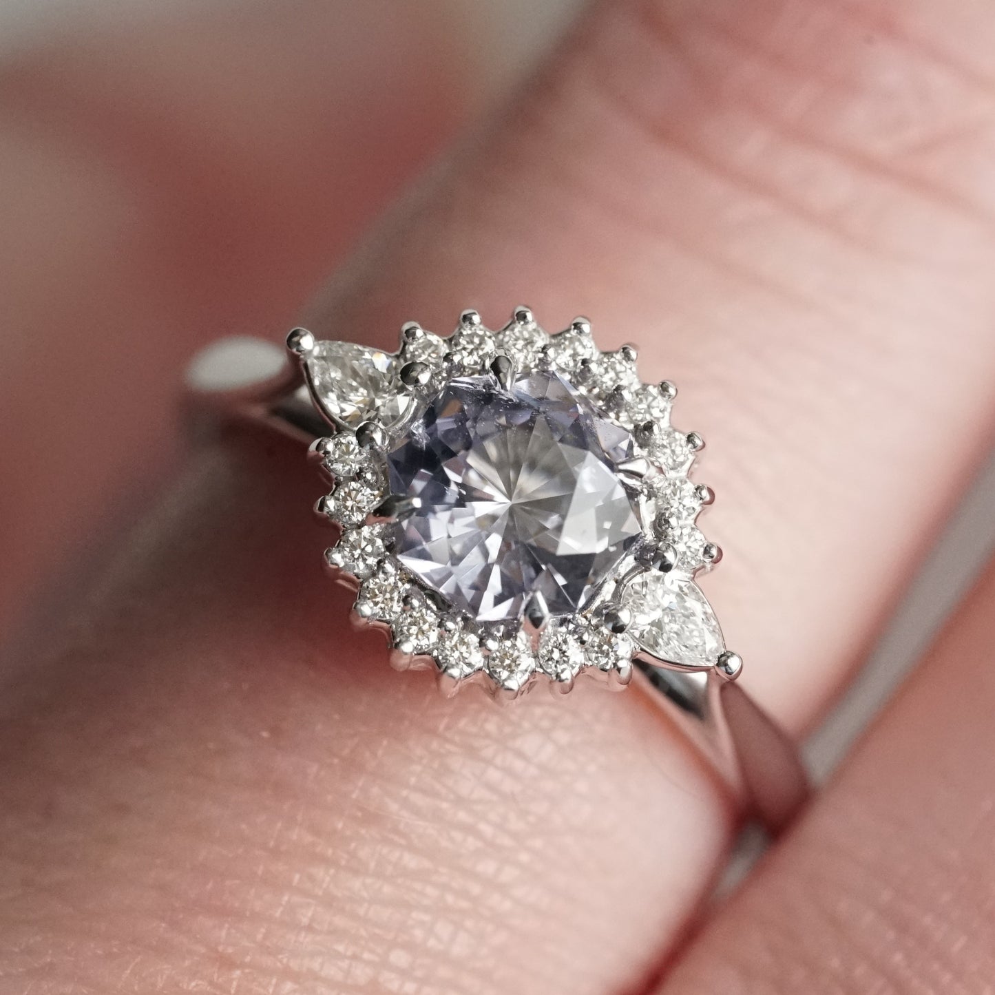 A macro photo of a lavender spinel and diamond halo style ring on a light-skinned left hand