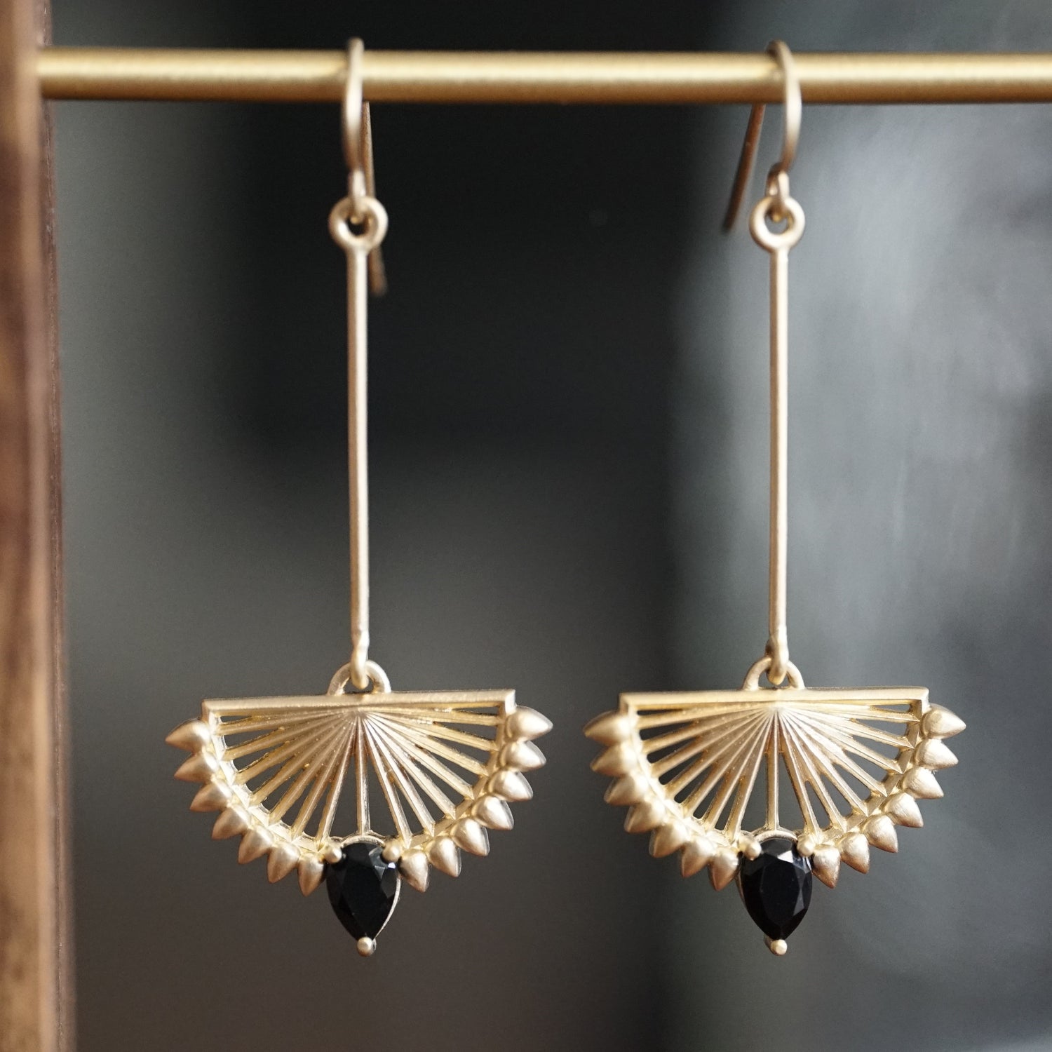 A pair of deco inspired matte finish yellow gold drop earrings with pear shape black onyx at the base hang on a brass wire