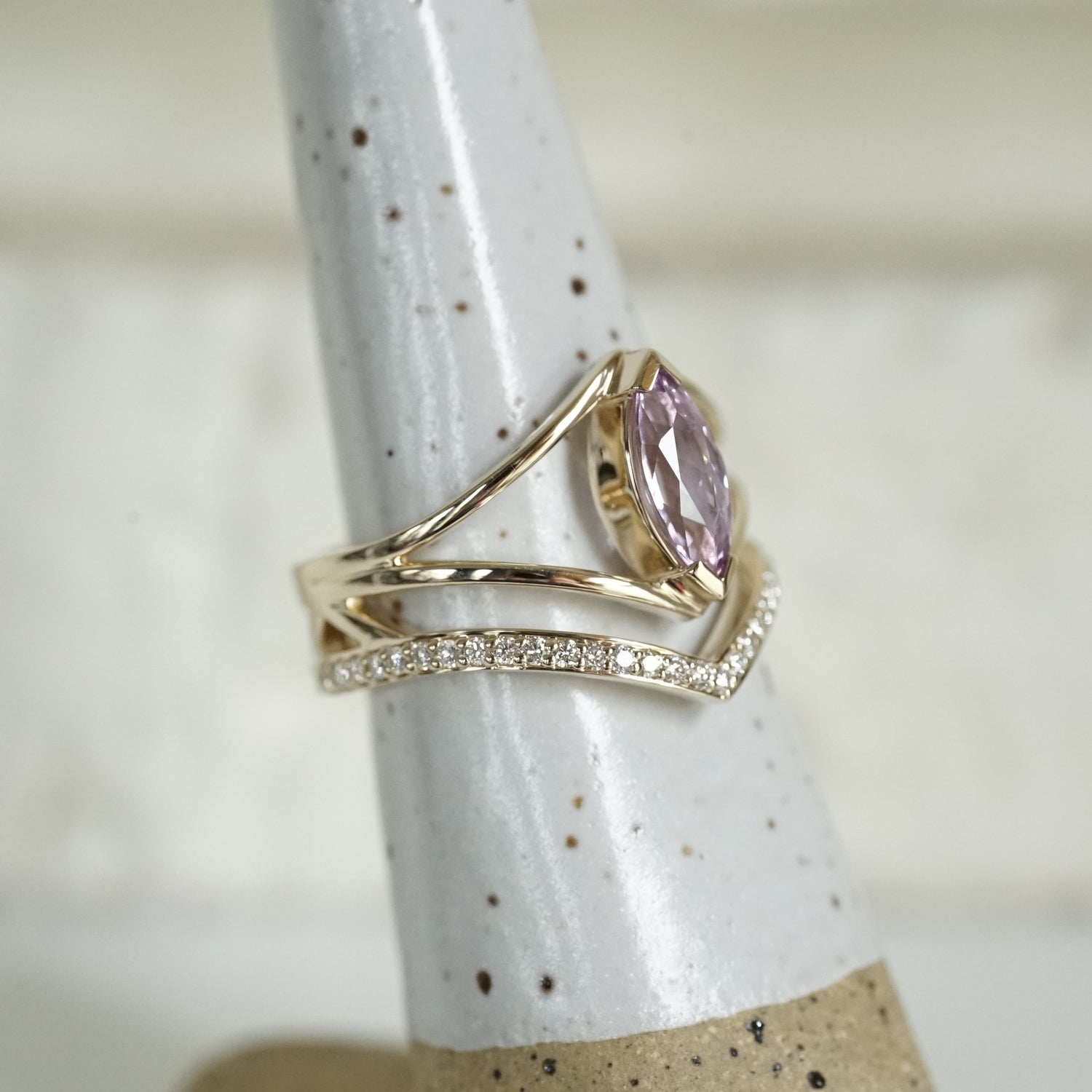 a side view of a yellow gold ring with a marquise cut lavender spinel between two V shape shadow style bands next to a V shape pave set diamond wedding band shown on a white ceramic display