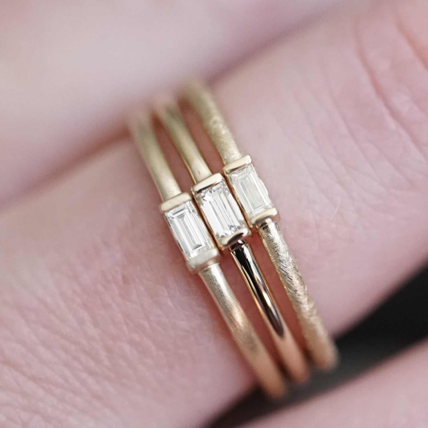 A set of 3 yellow gold stacking bands with various metal texture finishes featuring one baguette diamond each held in a light-skinned left hand