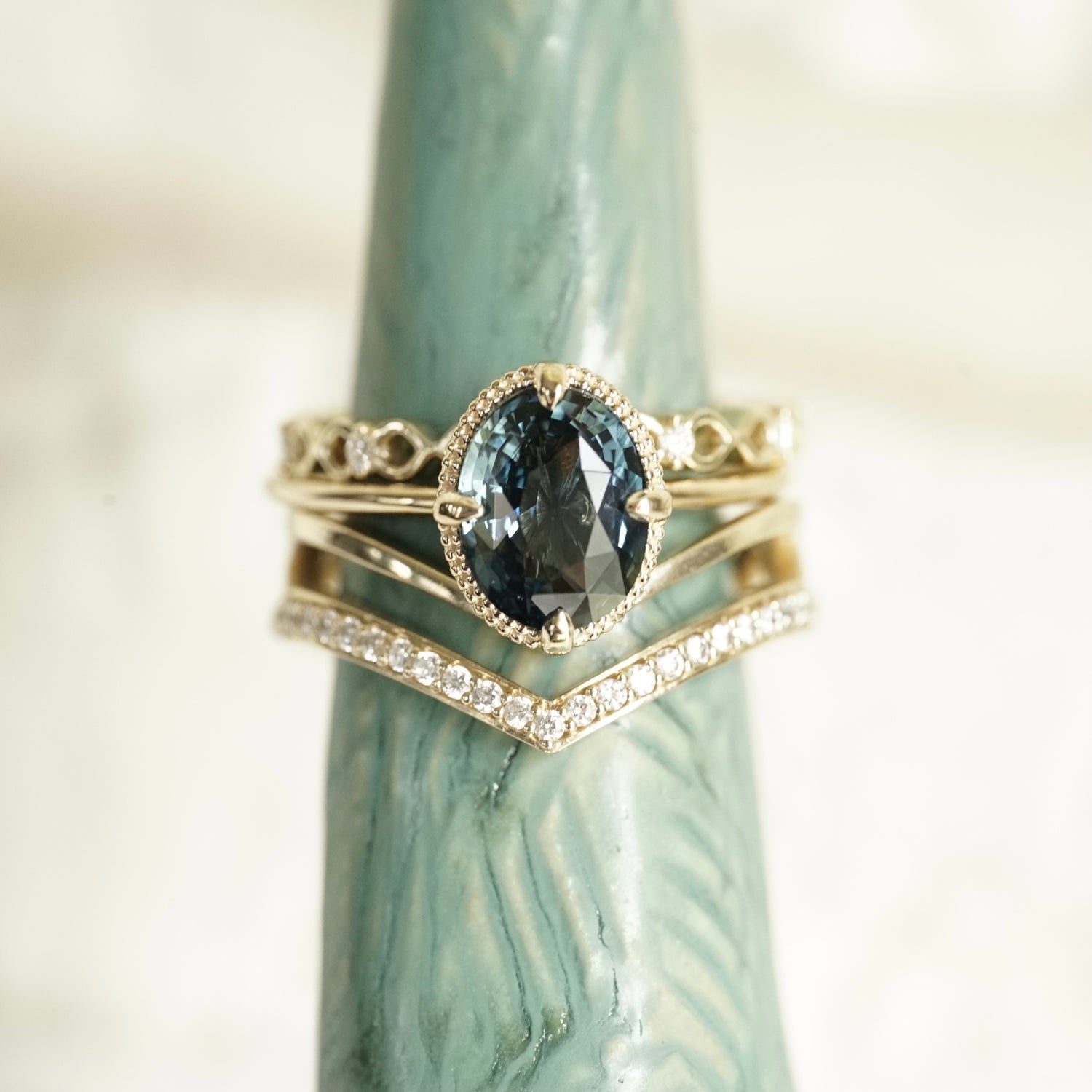 an oval teal sapphire is set in a yellow gold ring with a beaded detail behind the stone. The ring is show stacked with 2 yellow gold and diamond wedding bands. The set is shown on a light teal ceramic display