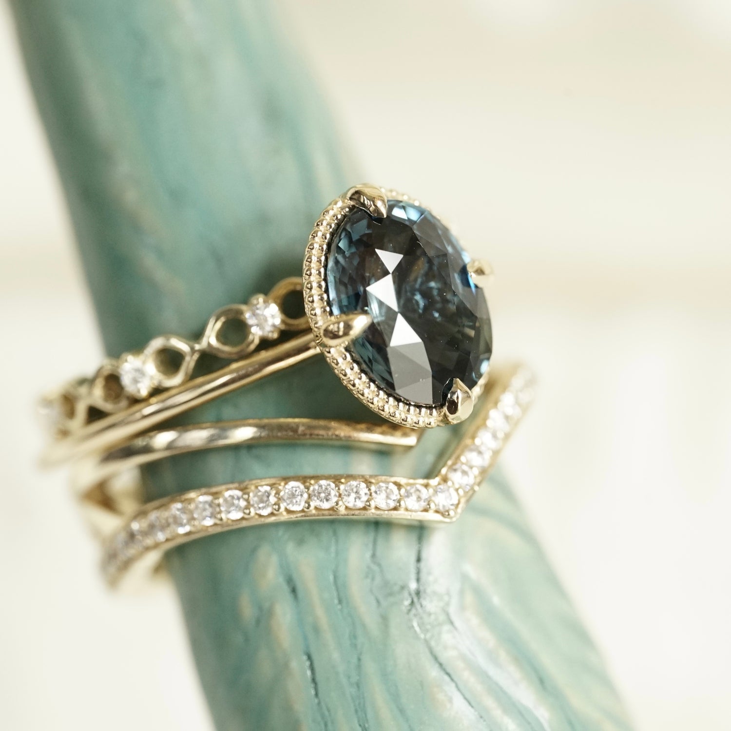 an oval teal sapphire is set in a yellow gold ring with a beaded detail behind the stone. The ring is show stacked with 2 yellow gold and diamond wedding bands. The set is shown on a light teal ceramic display