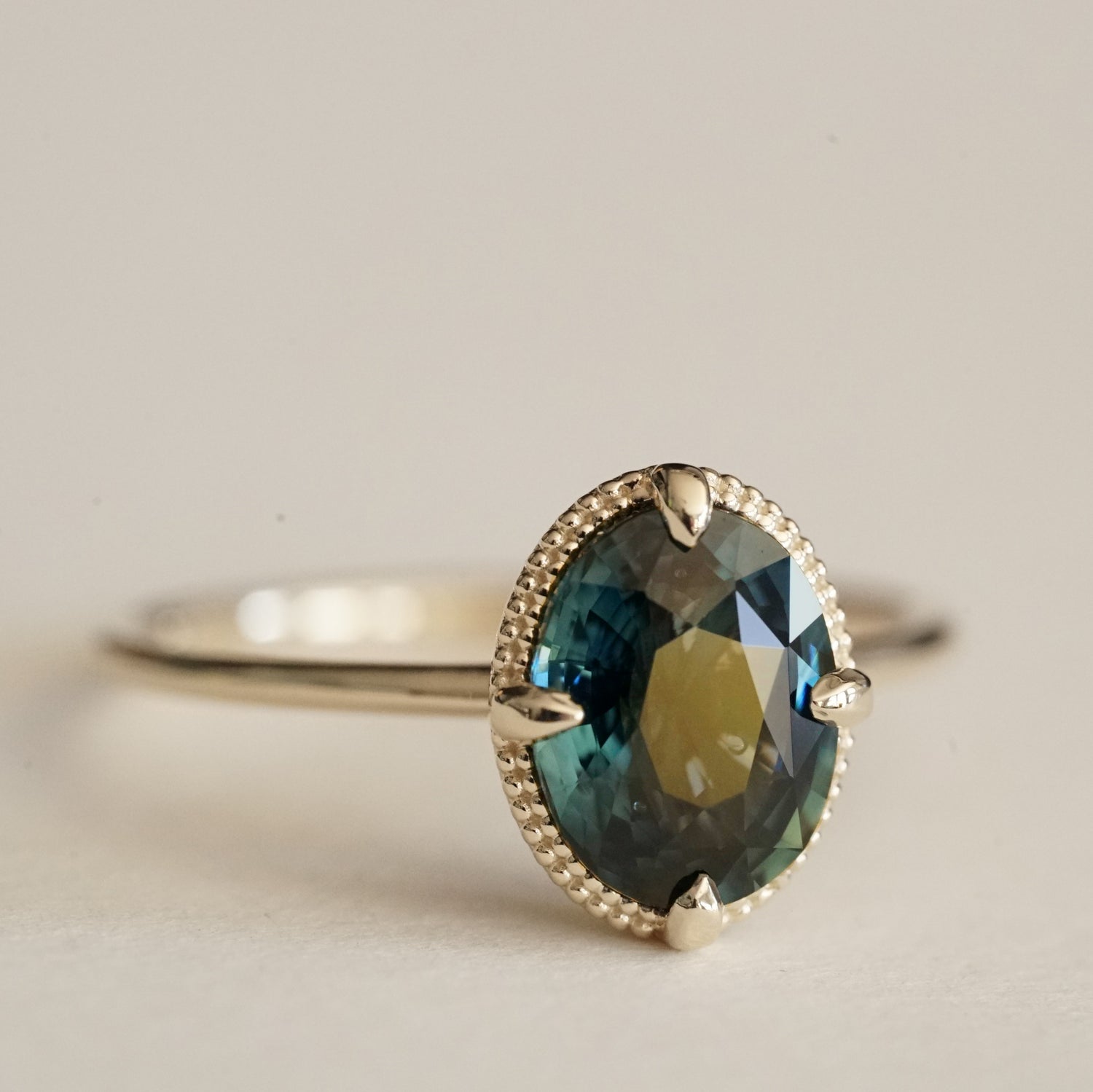 a macro photo of an oval teal sapphire is set in a yellow gold ring with a beaded detail behind the stone on a white matte background