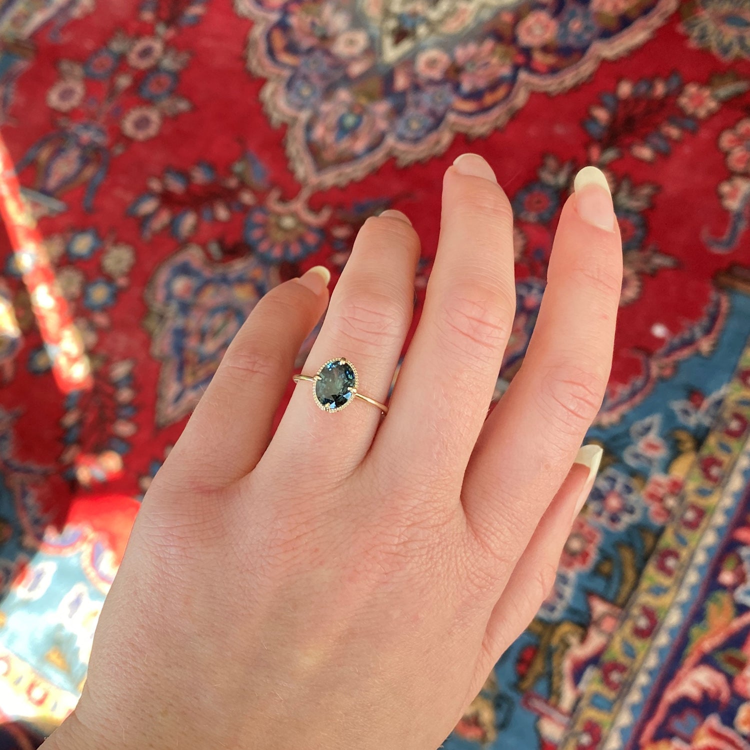 an oval teal sapphire is set in a yellow gold ring with a beaded detail behind the stone on a light skinned left hand