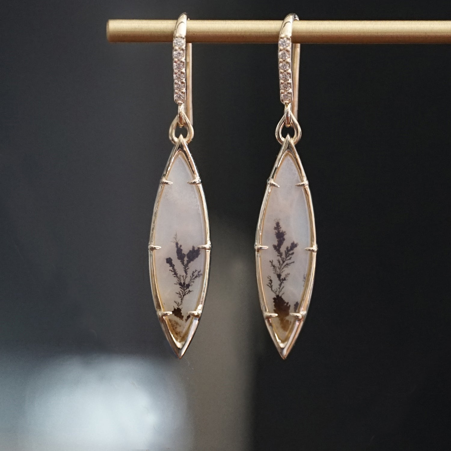 a pair of yellow gold drop earrings featuring dendritic agates and diamond drop tops hanging on a brass bar