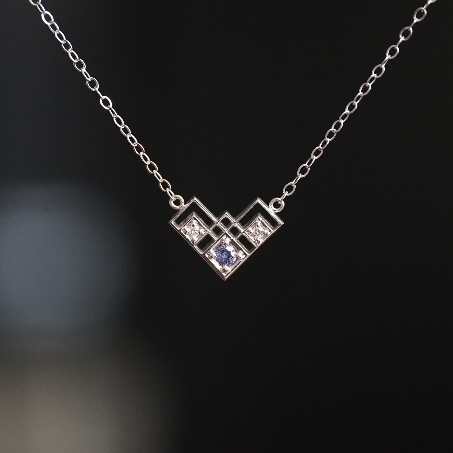 a white gold pendant with a light blue sapphire and 2 white diamonds in a semi heart shape art deco inspired pendant suspended on an open link white gold chain