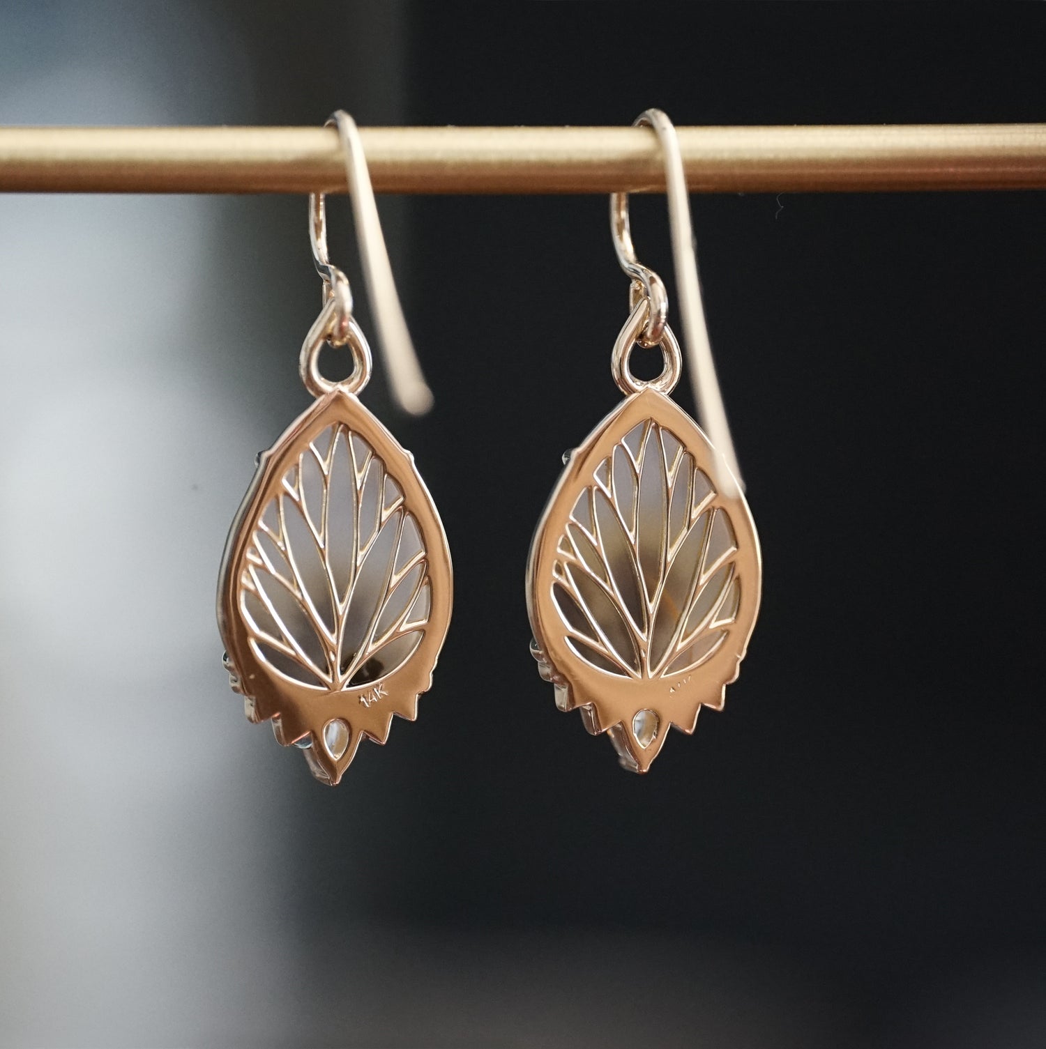 a pair of yellow gold drop earrings with dendritic agate center stones a single diamond accent at the base hang on a brass bar and are rotated to show the backside leaf detailing