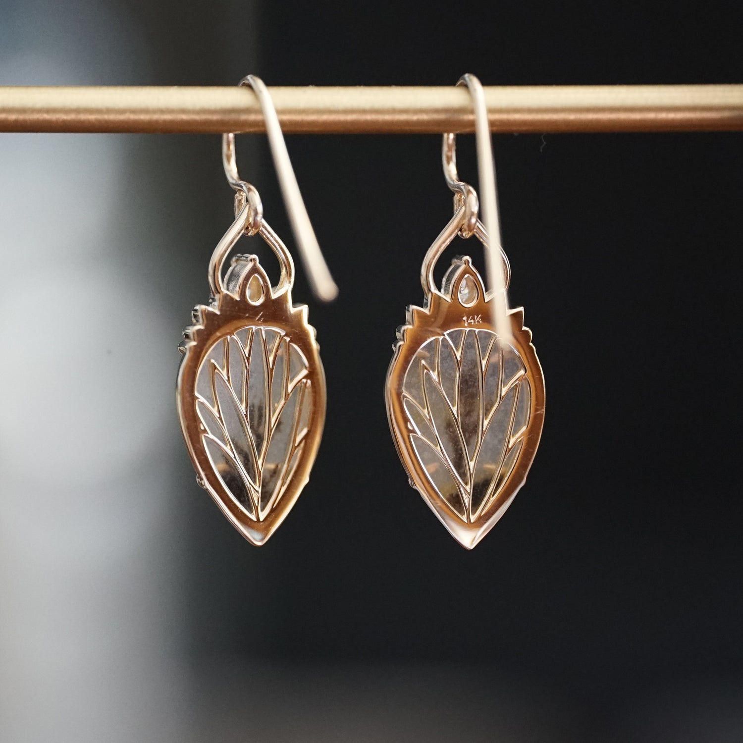 a pair of yellow gold drop earrings with dendritic agate center stones a single diamond accent at the top hang on a brass bar and are rotated to show the leaf detailing on the back