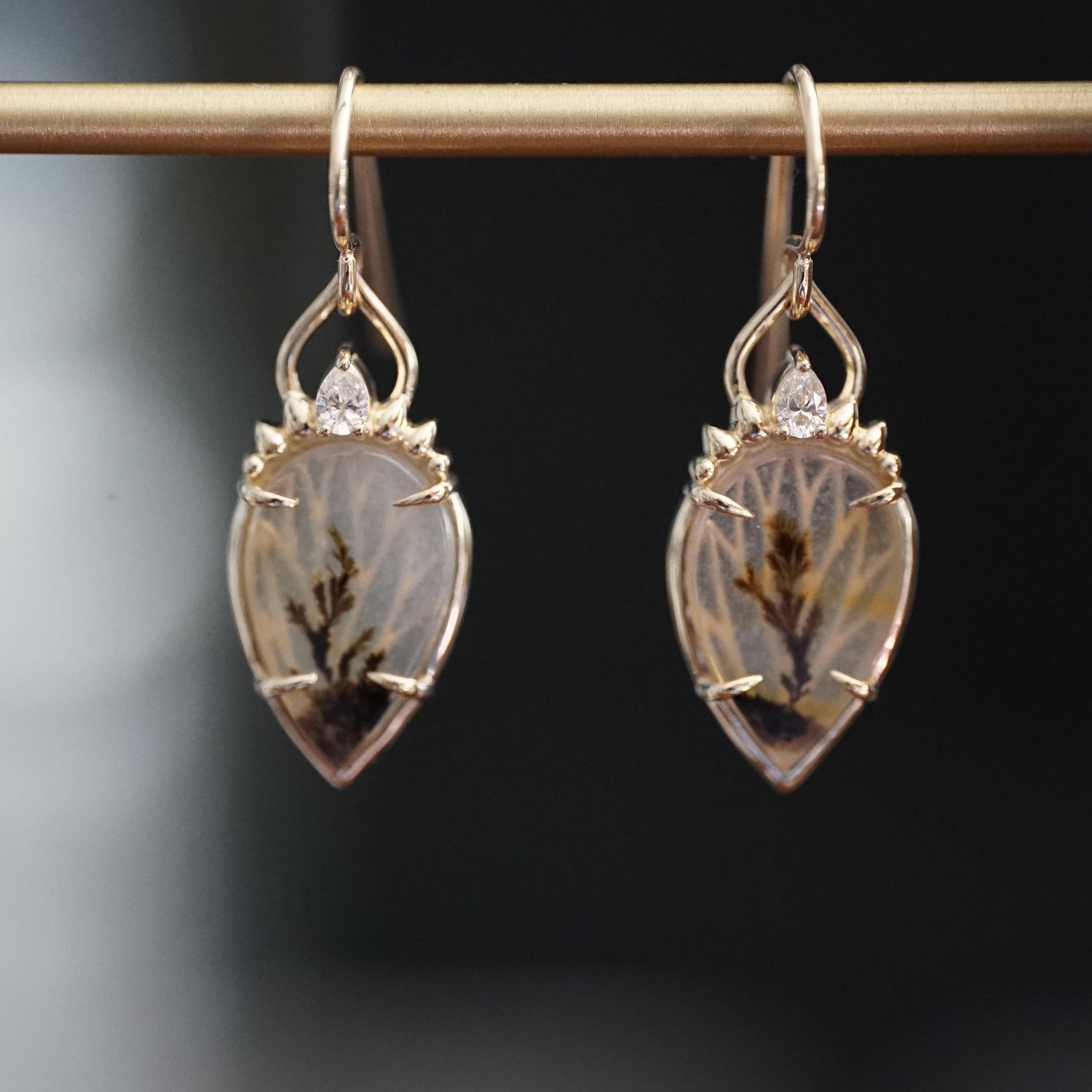 a pair of yellow gold drop earrings with dendritic agate center stones a single diamond accent at the top hang on a brass bar