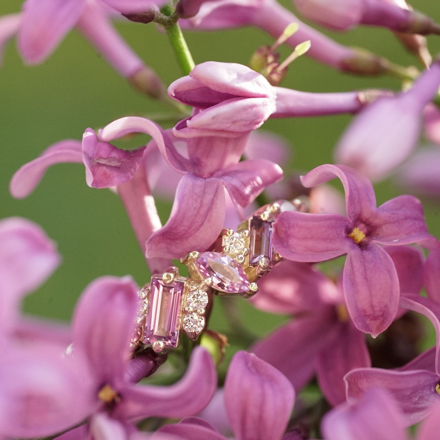 a yellow gold ring with pink sapphires of mixed shapes are paired with round white diamonds in a playful design rests in a bouquet of pink lilac flowers