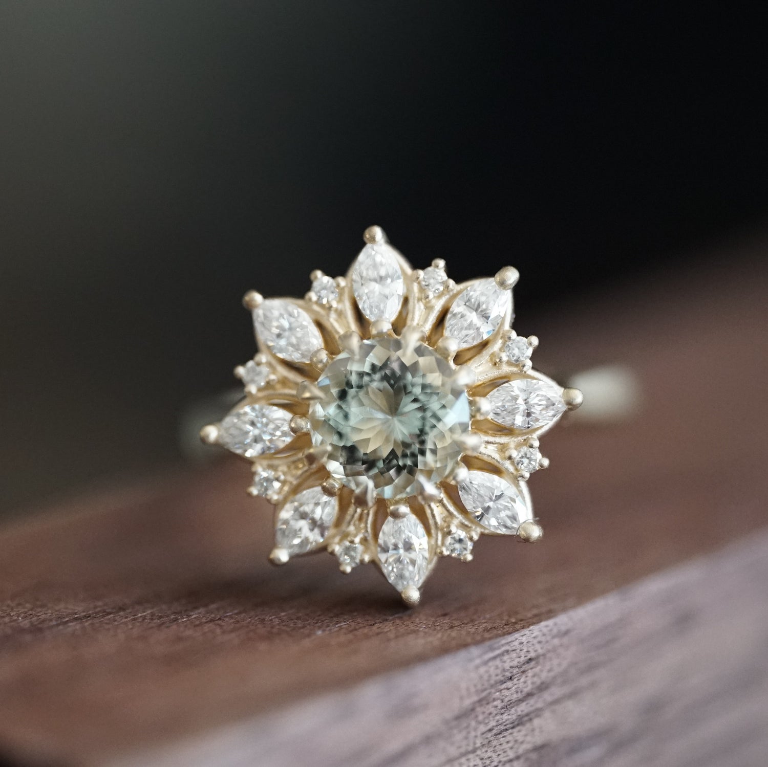 a yellow gold ring with a very light green prasiolite center stone and mixed gem diamond halo is resting on a wood display