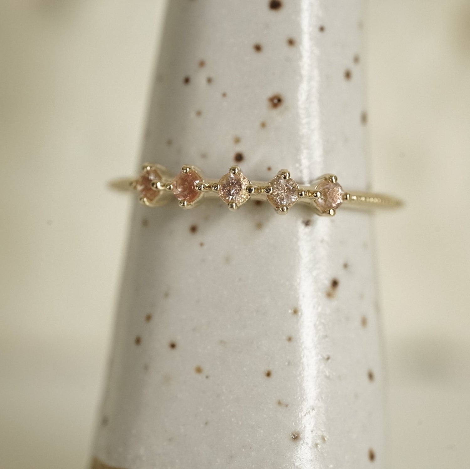 a yellow gold ring with 5 round light pink Oregon sunstones and migraine detail on the shank on a white ceramic display