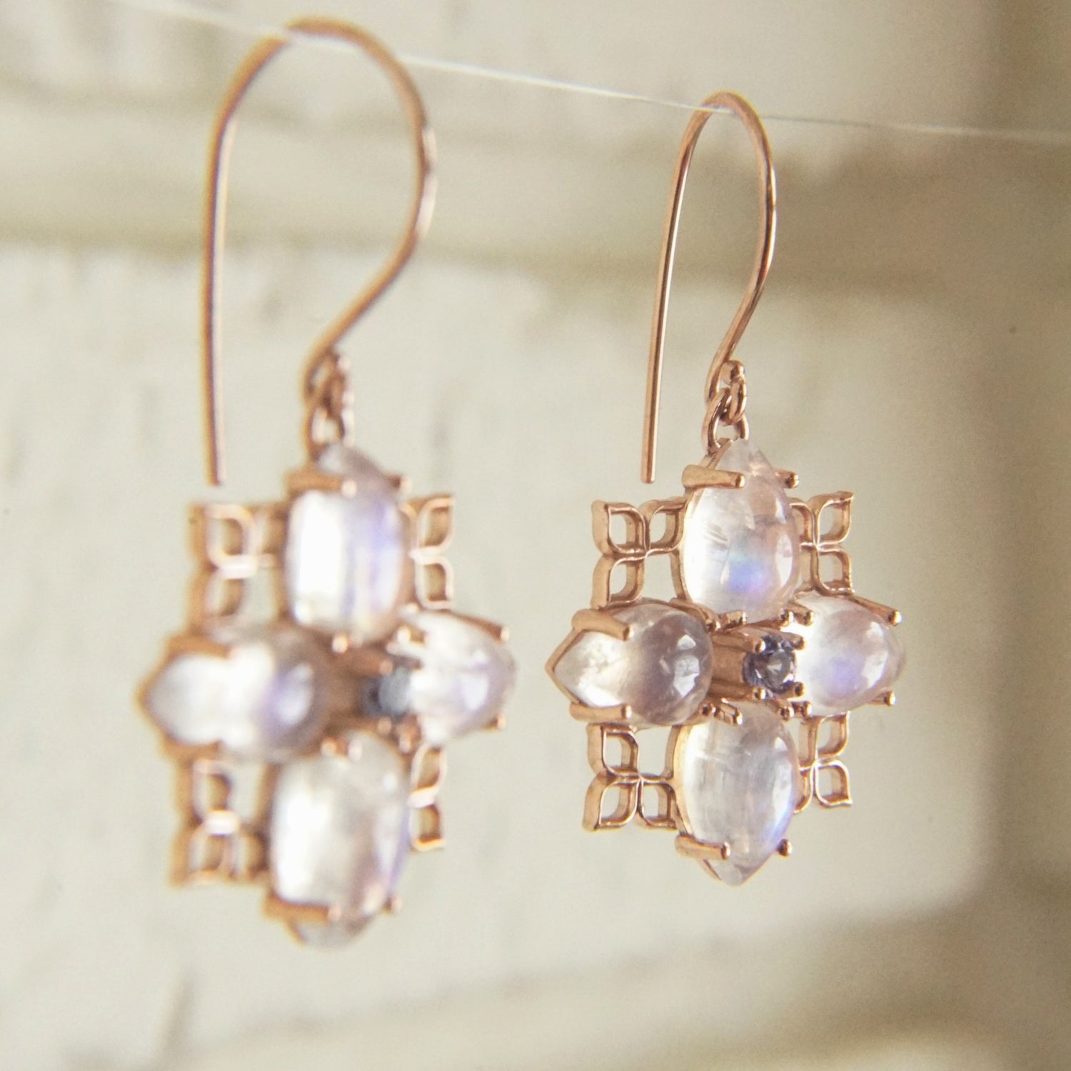 A pair of 14K Rose Gold, purple sapphire, and rainbow moonstone drop earrings hanging on a thin wire in front of a white brick wall