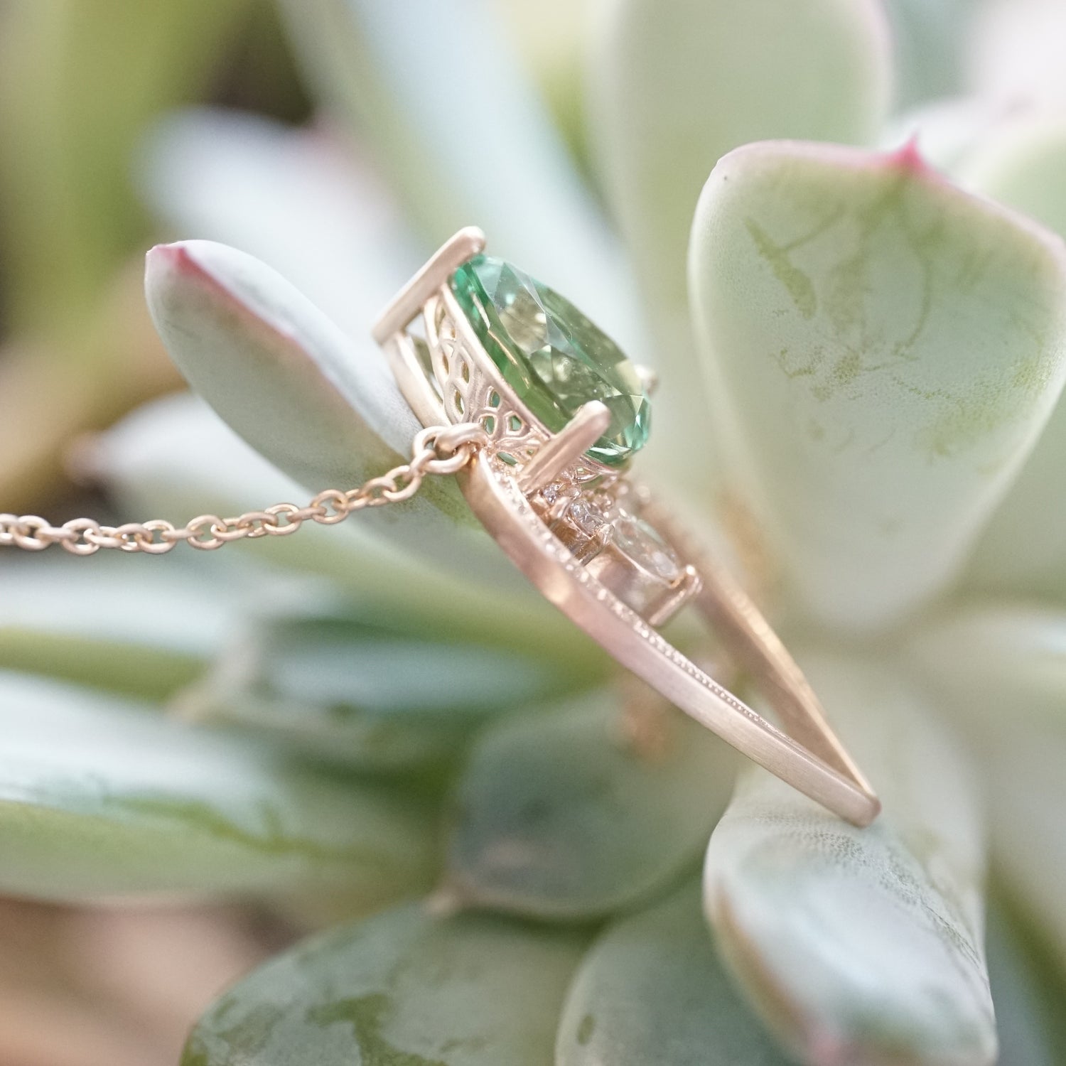 side view of a yellow gold pendant with a vibrant green tourmaline pear shape and diamond accents suspended on a cable chain and laying on a succulent