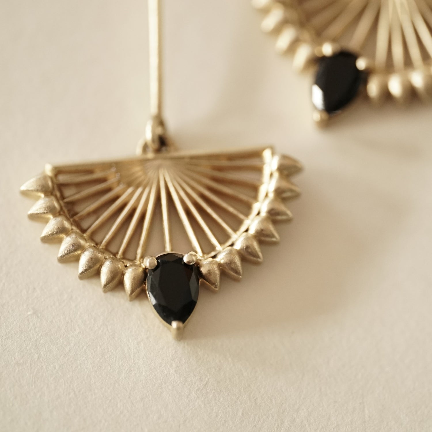 A macro photo of a pair of deco inspired matte finish yellow gold drop earrings with pear shape black onyx on a matte white background
