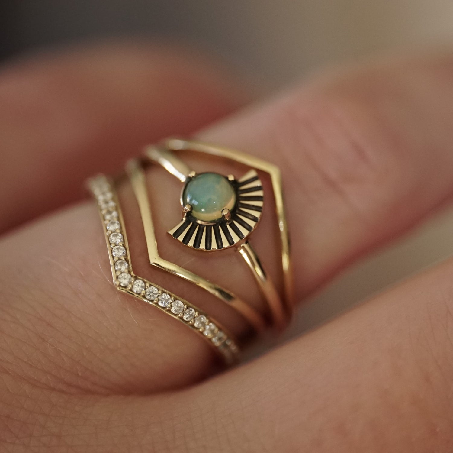 a cabochon cut opal ring with deco fan element to one side is stacked with v shape shadow bands, one with diamonds and 2 without on a light-skinned left hand