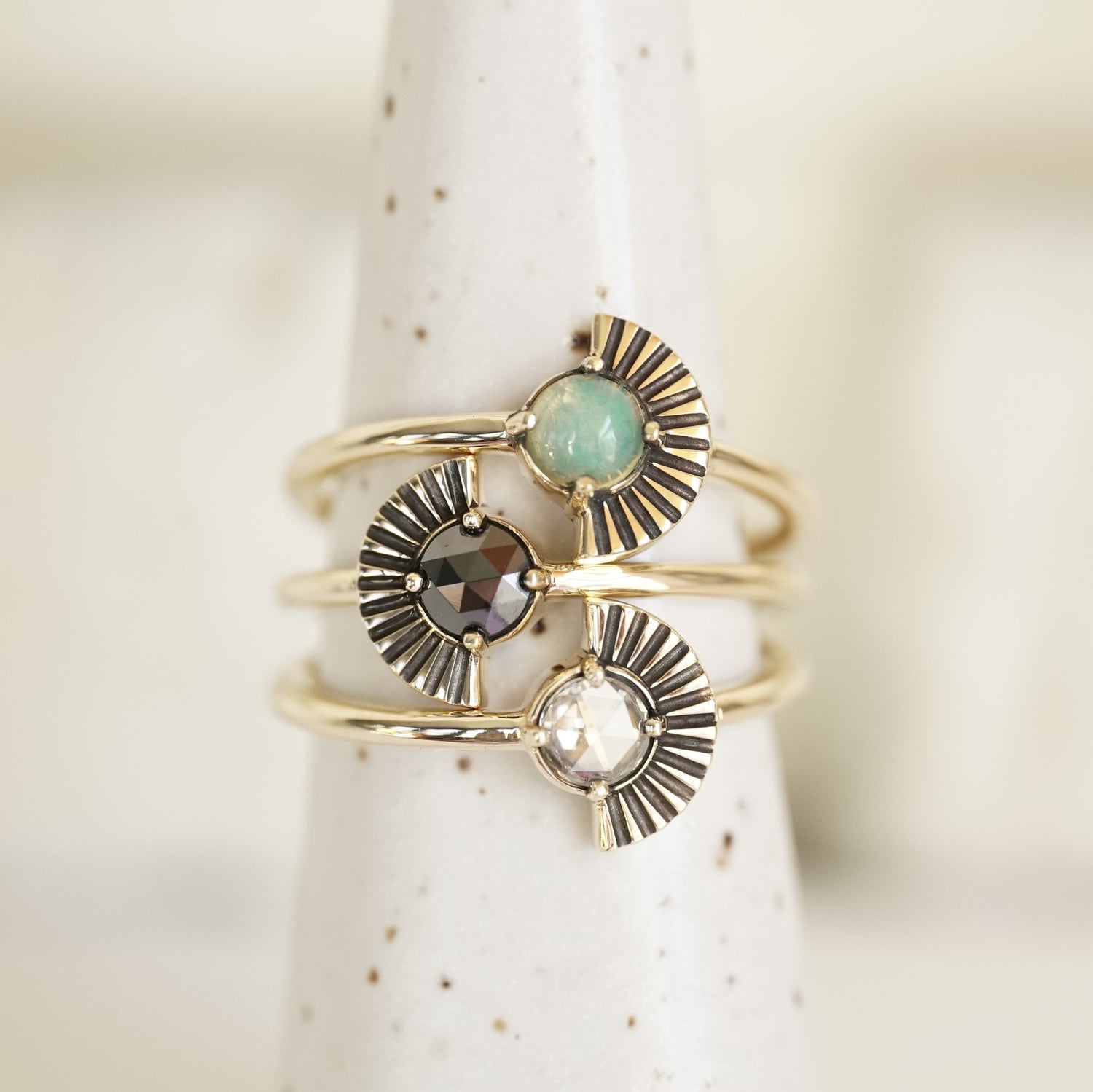 a set of 3 yellow gold deco inspired rings each featuring a single round gem, one opal, one black diamond, and one white diamond. The gems are prong set and there is a fan shape element to one side. the rings are on a white ceramic display