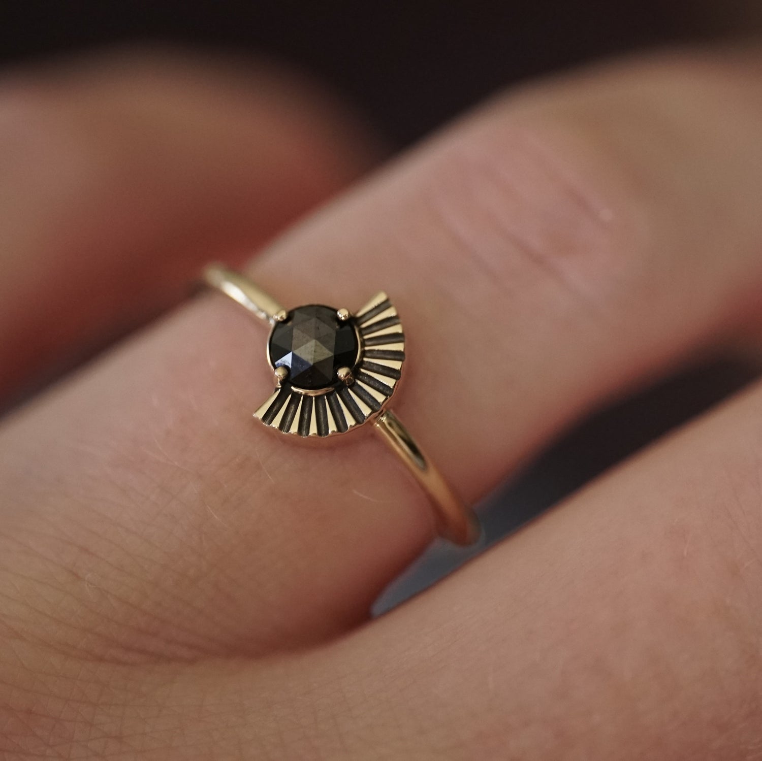 a rose cut black diamond in a yellow gold ring with an art deco fan element to one side is shown on a light-skinned left hand