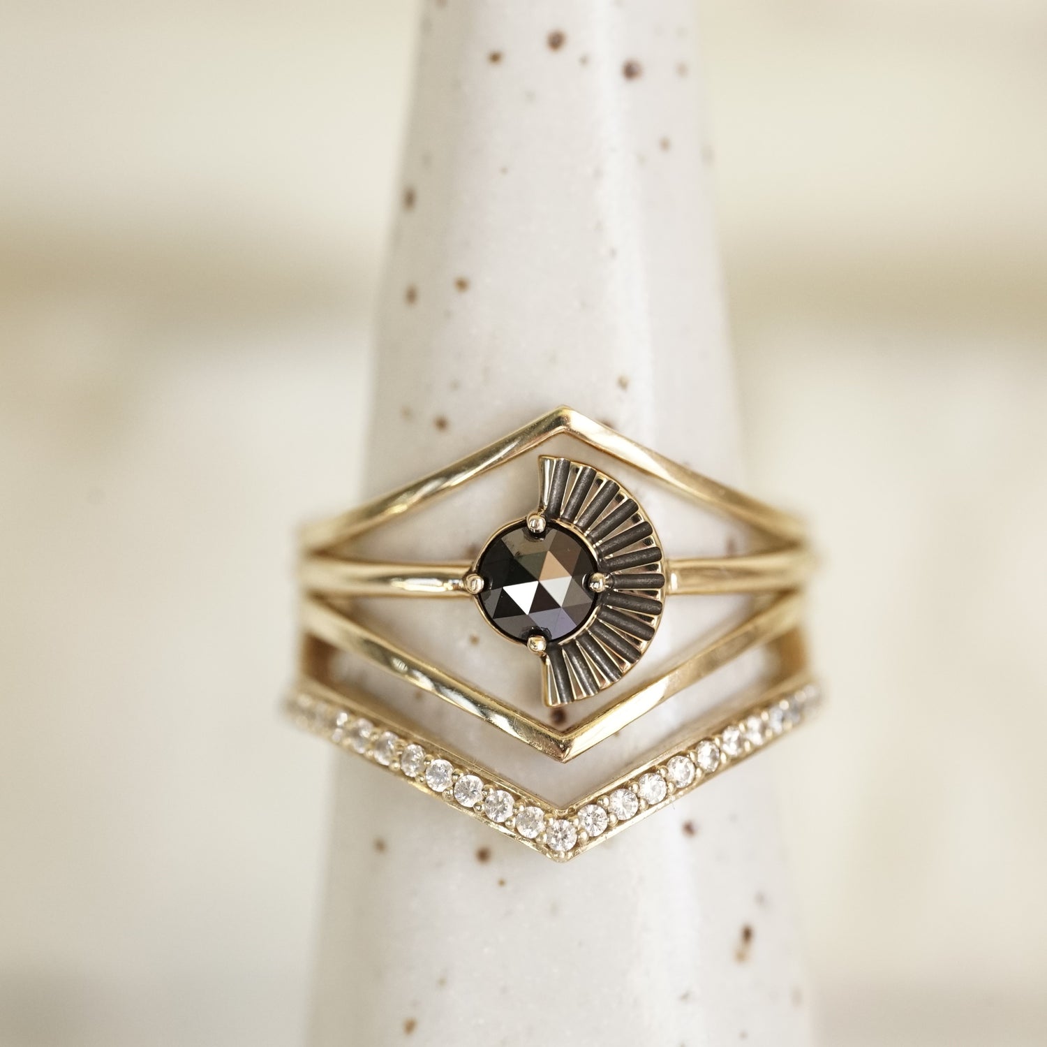 a rose cut black diamond ring with deco fan element to one side is stacked with v shape shadow bands, one with diamonds and 2 without on a white ceramic display