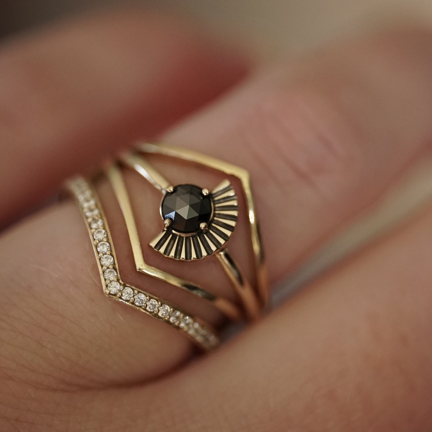a rose cut black diamond ring with deco fan element to one side is stacked with v shape shadow bands, one with diamonds and 2 without on a light-skinned left hand