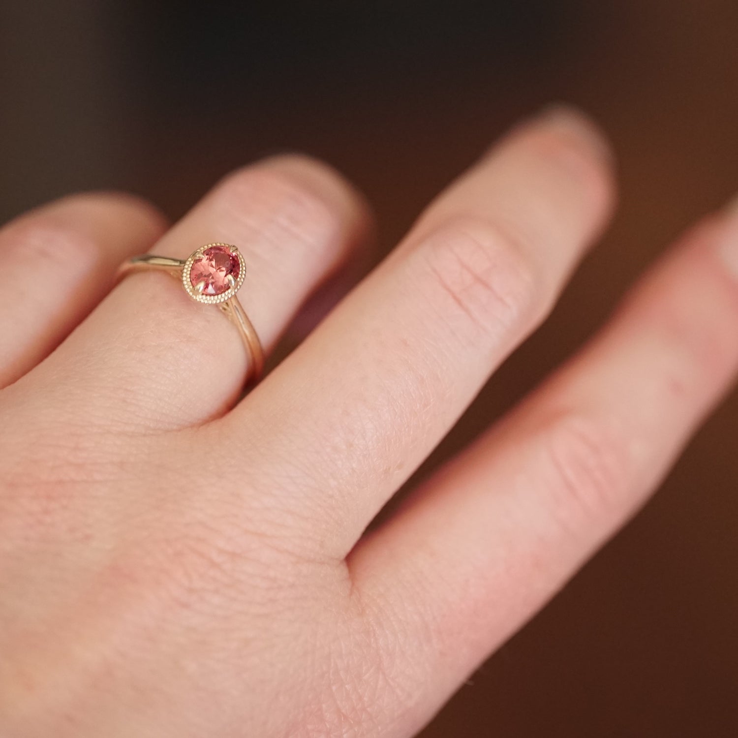 a macro photo of a vibrant oval pink spinel in a yellow gold ring setting with a beaded halo on a light skinned left hand