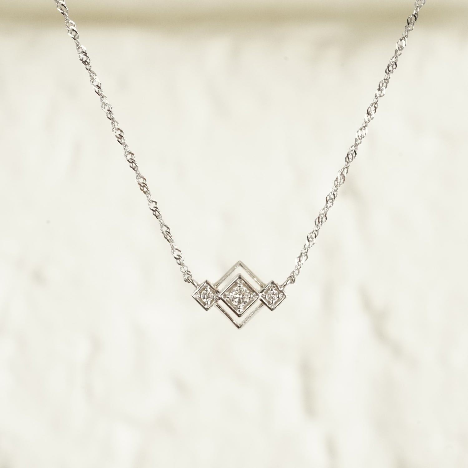 an art deco inspired white gold pendant with 3 round diamonds in squares on point attached to a sparkly white gold chain