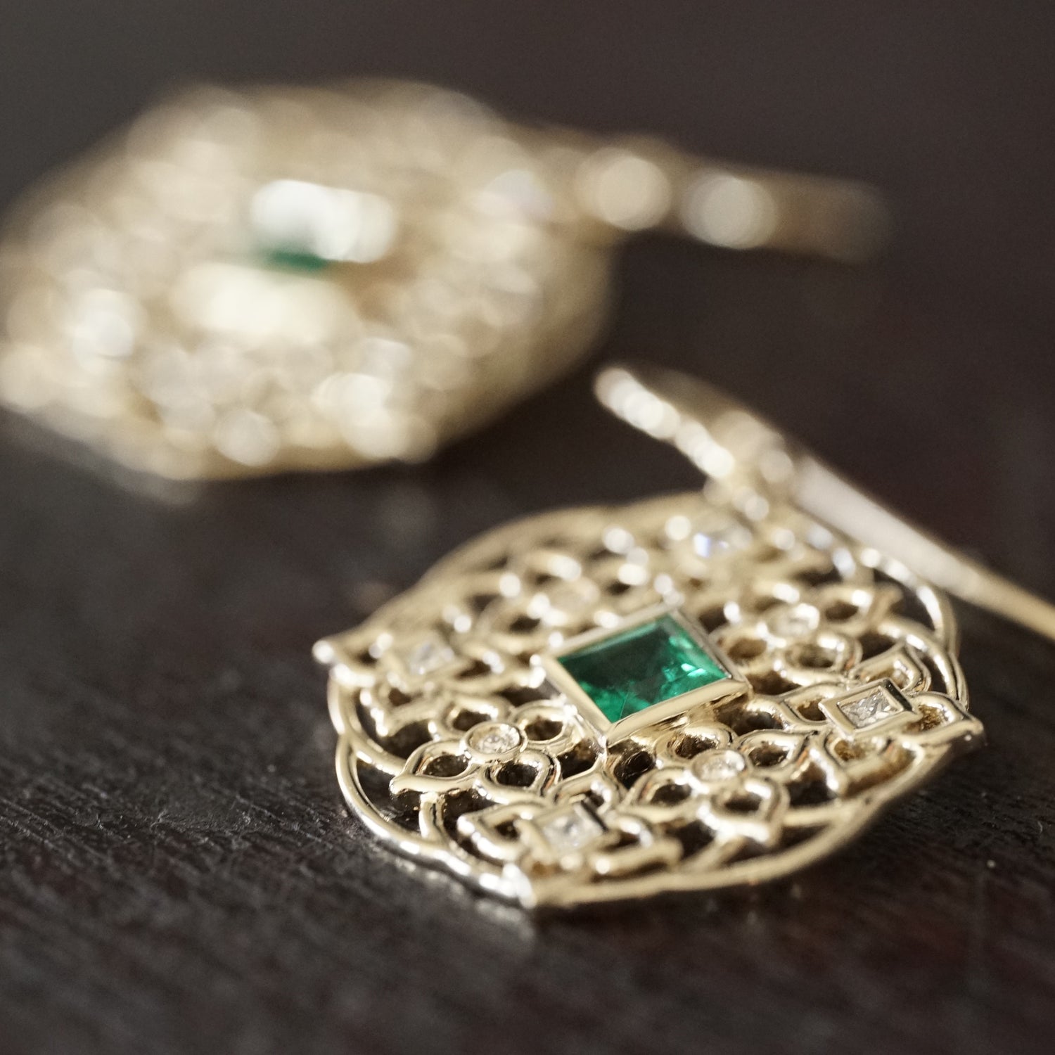 a macro photo of a yellow gold earring with princess cut emerald center stones and round and princess cut diamond earrings rests on a wood background