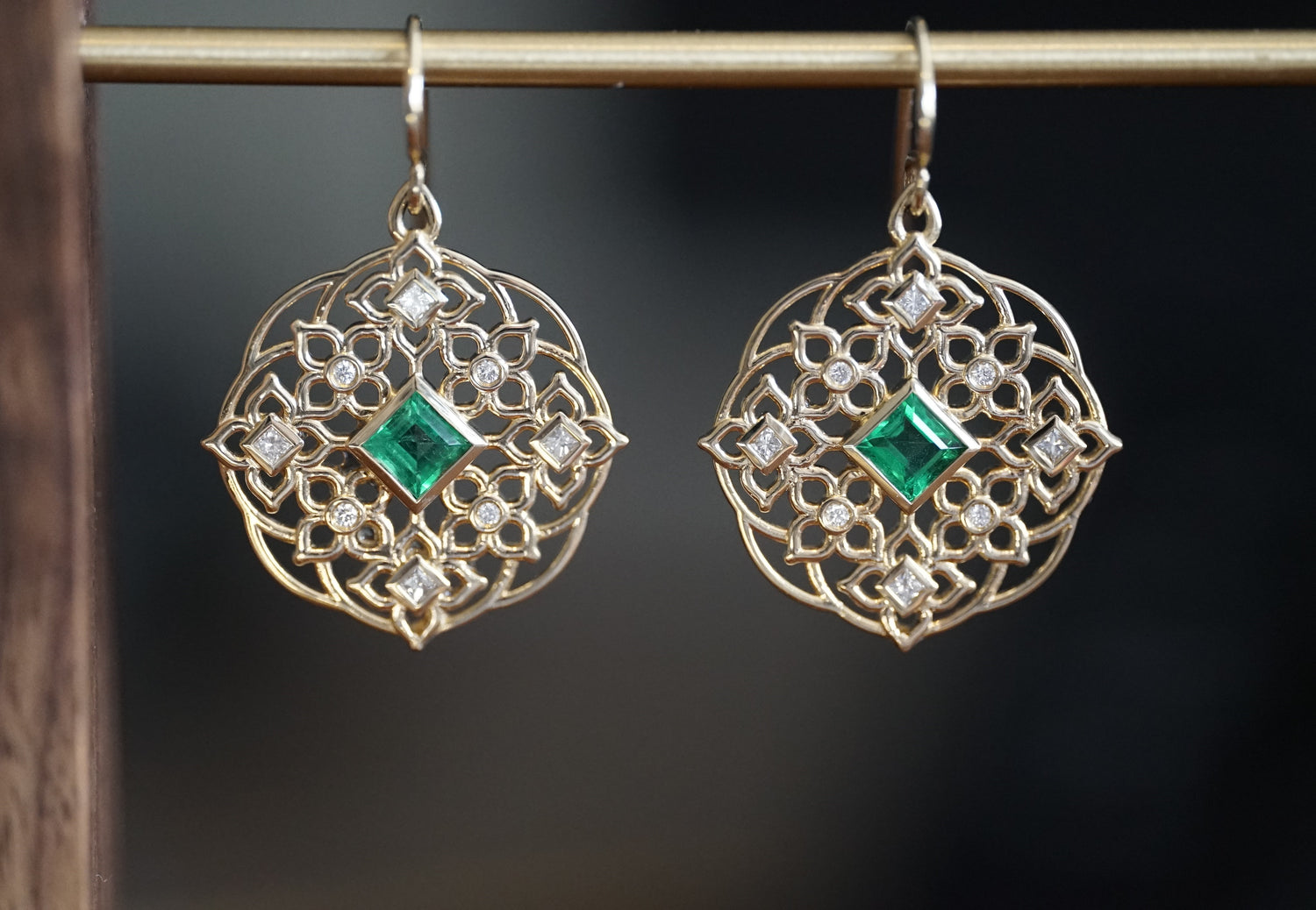 a pair of yellow gold earrings with princess cut emerald center stones and round and princess cut diamond earrings hang on a brass bar