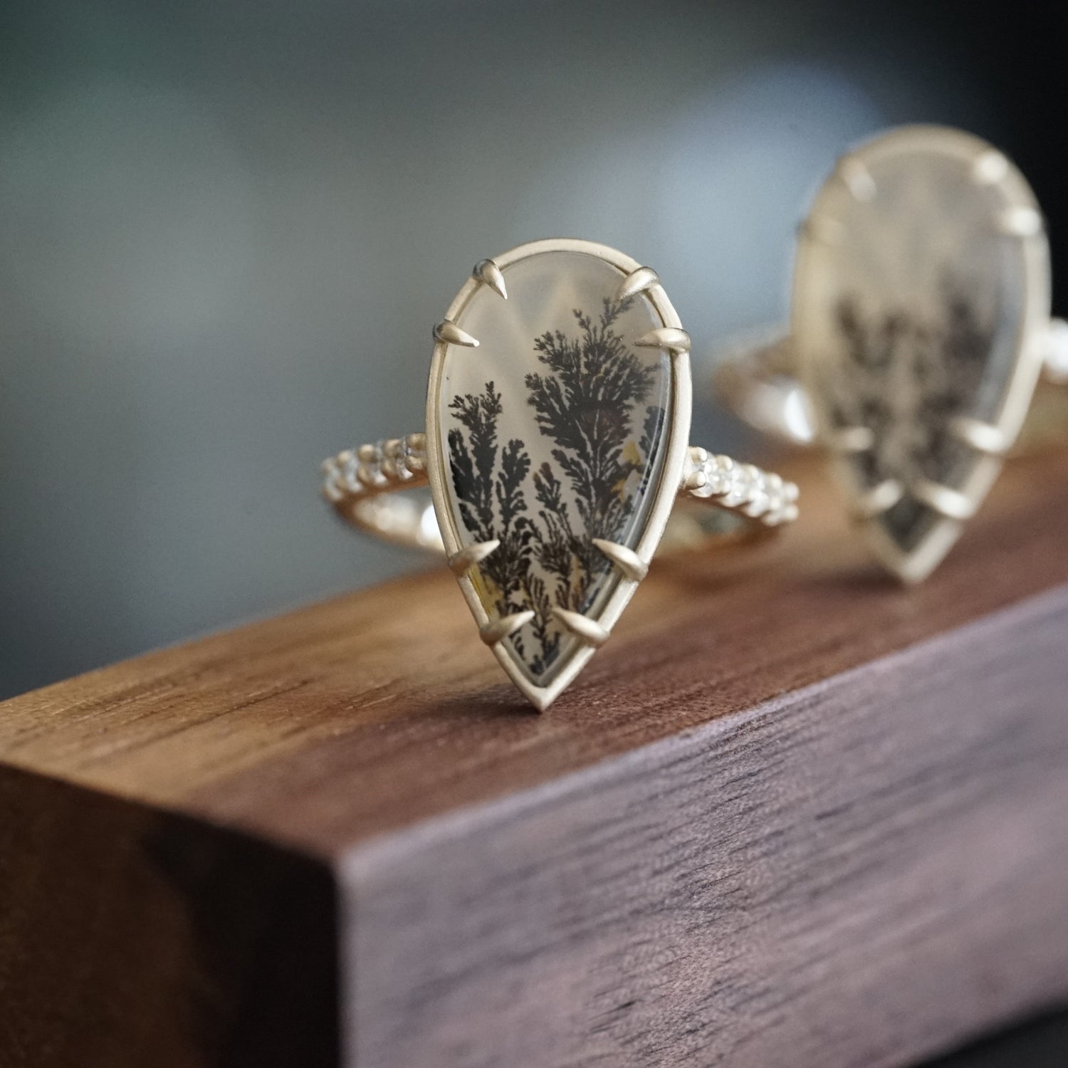 a yellow gold ring featuring a pear shape dendritic agave and shared prong diamonds on the shank rests on a wood display